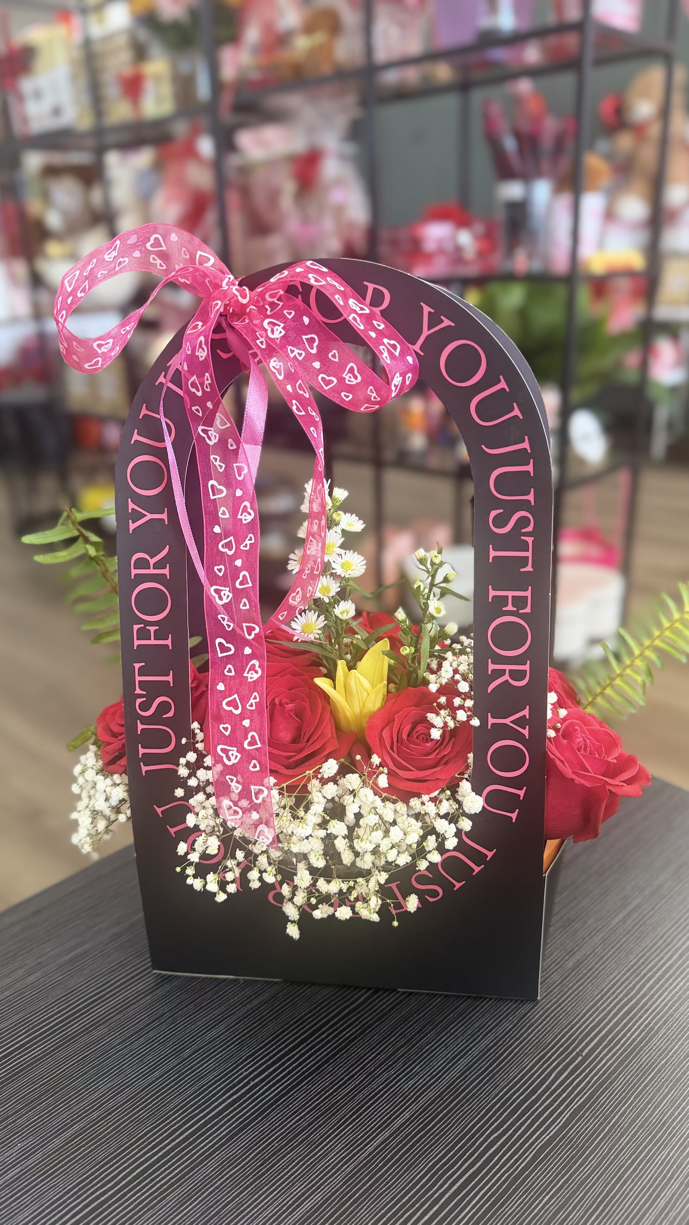 Pink roses and a yellow lily in a black gift bag with a heart ribbon.