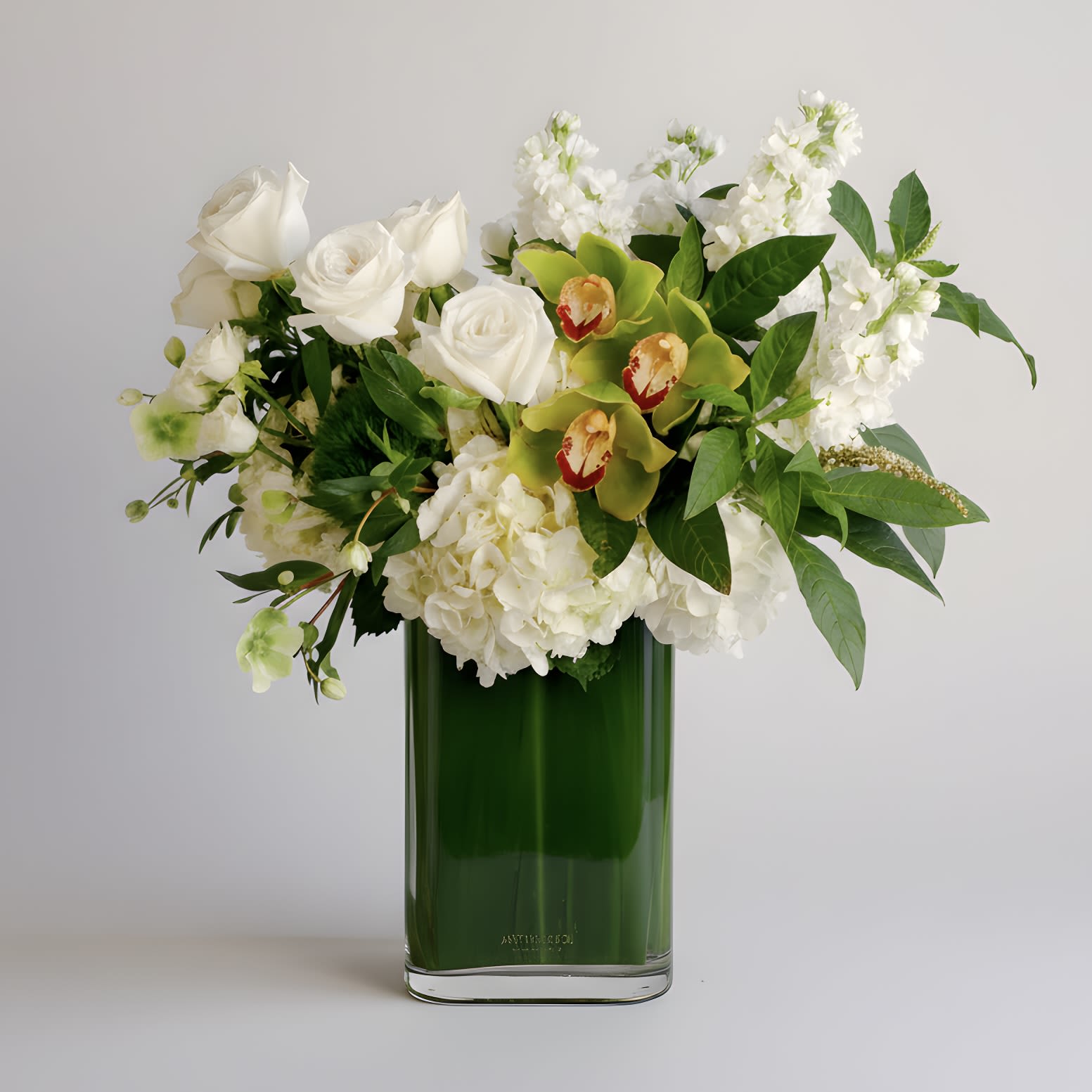 Emerald Elegance - This modern arrangement features an elegant collection of green cymbidium orchids, white roses, hydrangeas, hellebores, fragrant stock and more in a tall rectangular leaf-lined glass vase. Approximately measures 20 inches tall and 18 inches wide with flowers.