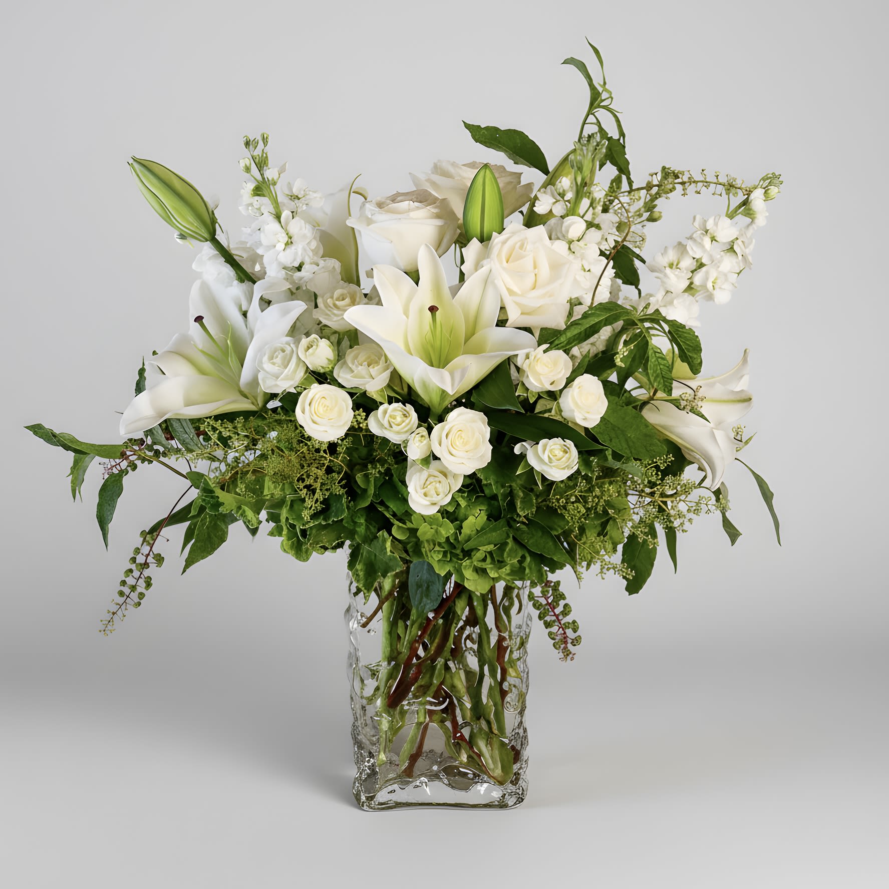 Elegant Blooms - This serene arrangement highlights premium white lilies mixed with white roses, spray roses, fragrant stock, green hydrangea and lush greenery in a unique glass vase. Arrangement approximately measures 18 inches tall and 12 inches wide with flowers.