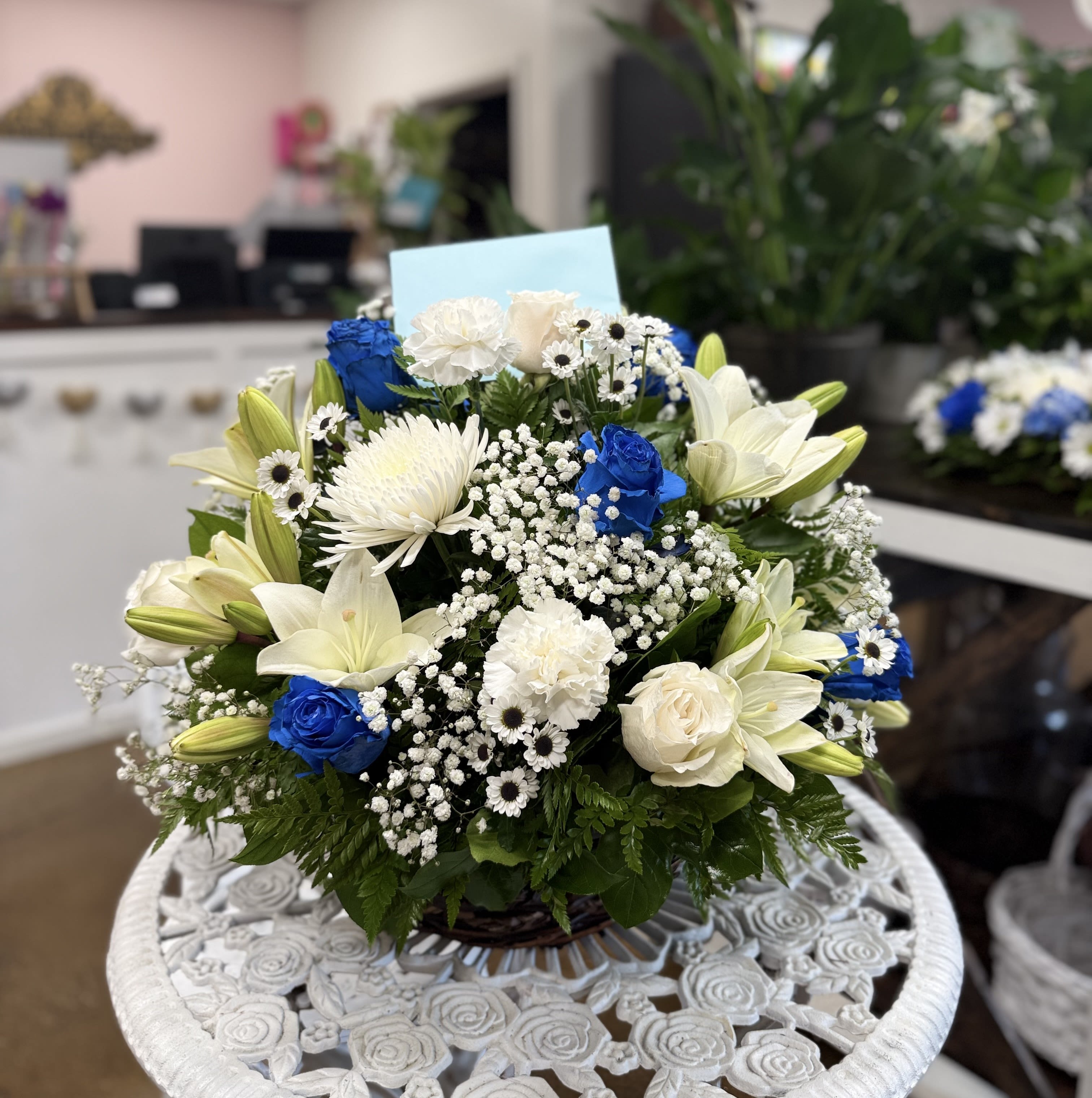 PEACEFUL MEMORIES BASKET - This elegant basket arrangement features a serene blend of white lilies, carnations, roses, and mums, beautifully accented with striking blue roses and delicate baby’s breath. Designed with lush greenery, it offers a timeless expression of peace, respect, and remembrance—an ideal tribute to honor a loved one’s memory. Size: Medium approximately 18 inches wide.    