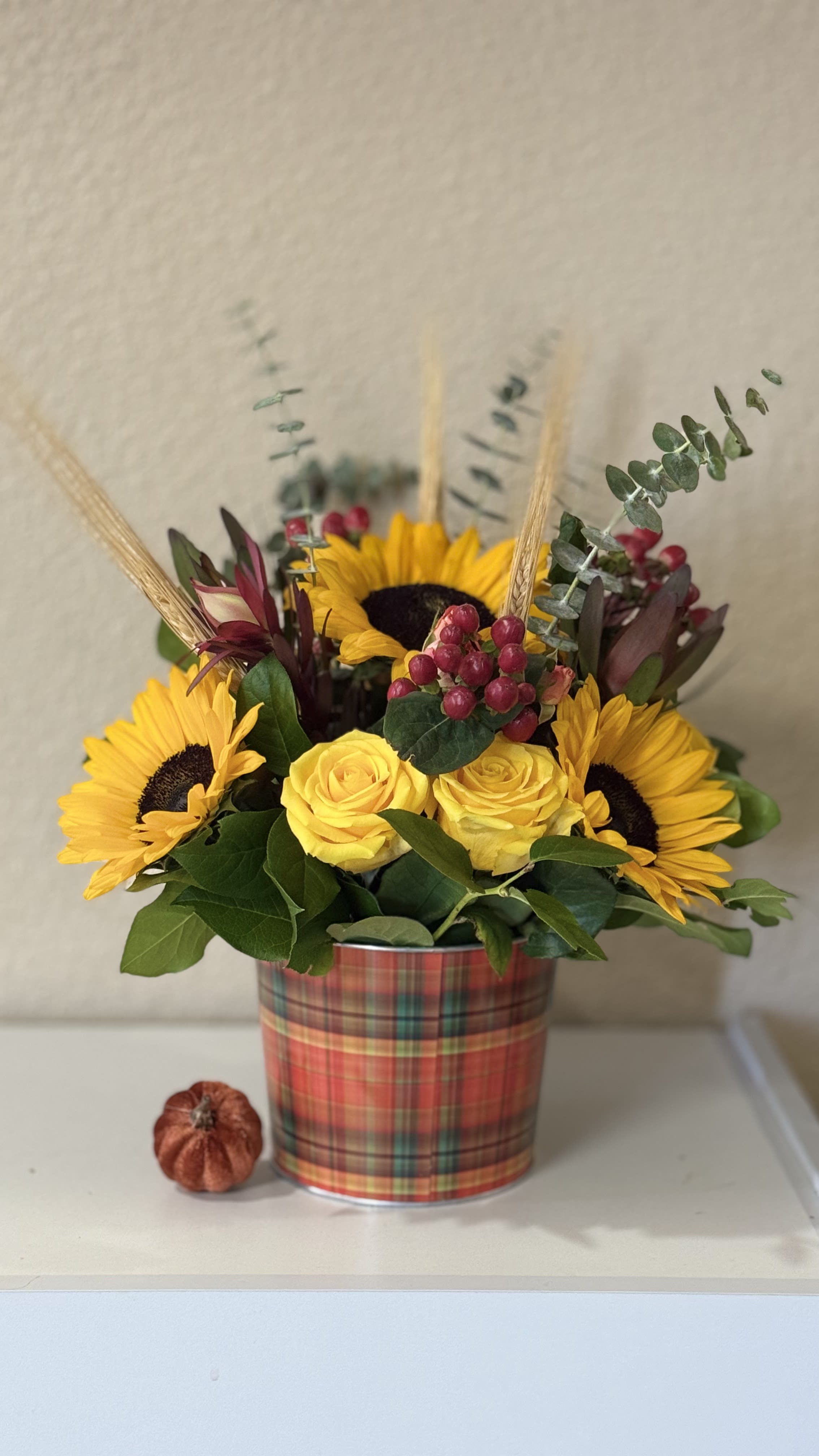 Golden Harvest Glow - Bright, warm, and full of joy this cheerful design brings together radian Sunflowers, golden roses, rustic hypericum berries, eucalyptus, all arranged in a cozy plaid container. It captures the heart  of autumn sunshine, gratitude, and nature's golden touch- perfect for Thanksgiving gatherings or brighten someone's day with a little happiness.