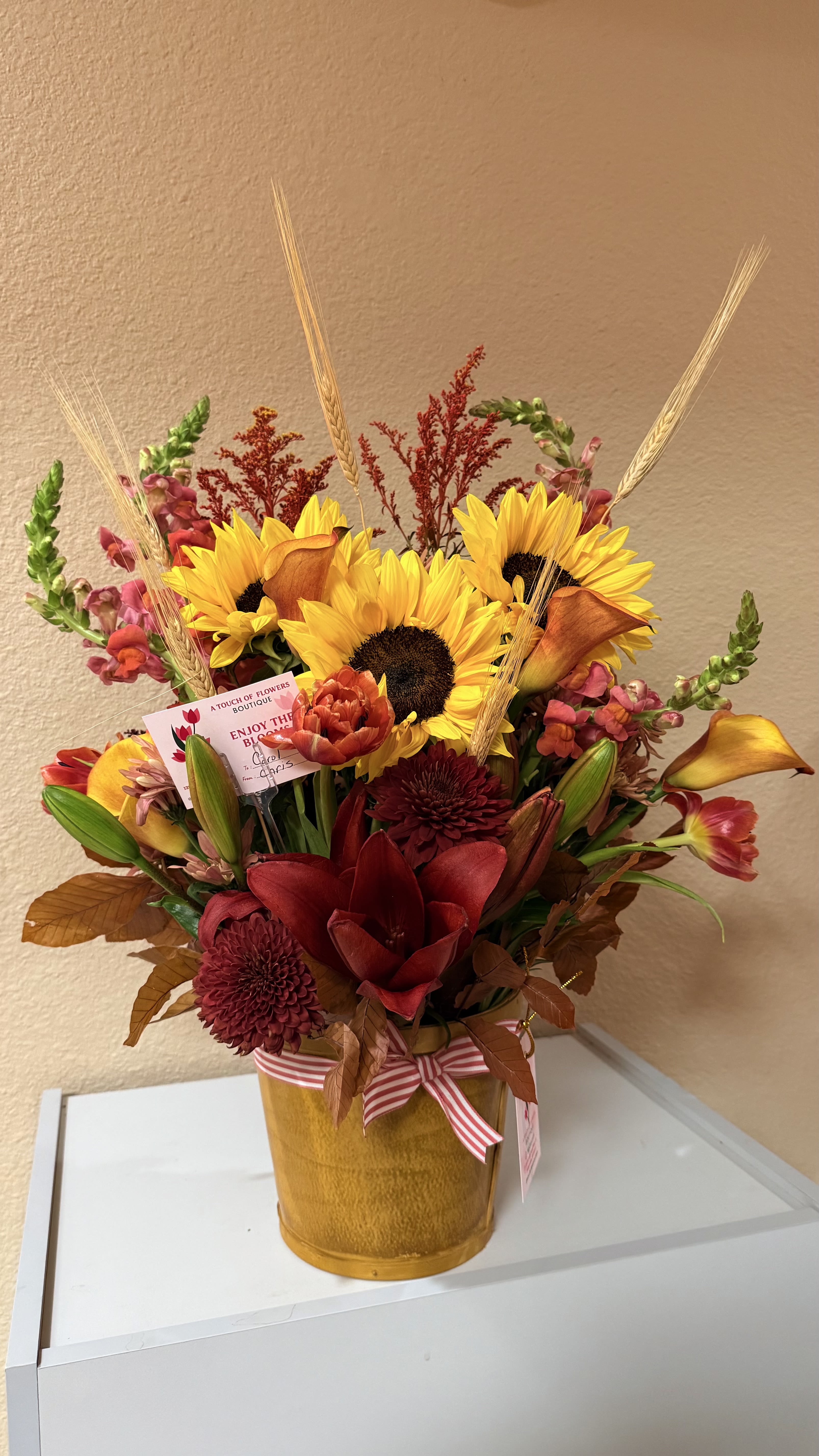 Golden Harvests - Radiating the rich  beauty of the fall season, brings together bright sunflowers, deep burgundy blooms, lilies, calla lilies, snapdragons, autumn leaves, and natural wheat stems. Designed in a rustic golden vase, this arrangement captures the heart of autumn _ warm inviting, and full of texture.