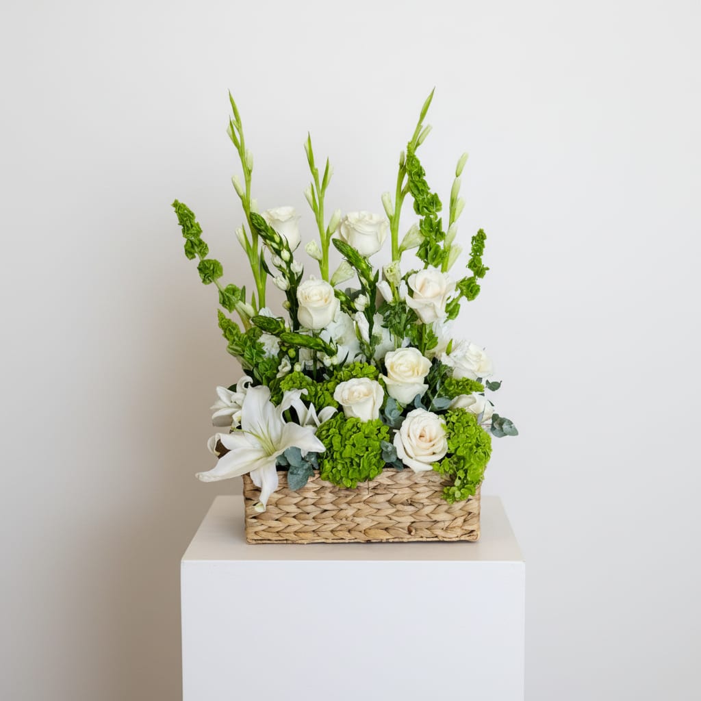 Eternal Grace Basket/ White Roses, Lilies and Bells of Ireland  - Pure white roses and open white lilies rise from a low woven rectangular basket, creating a calm, elegant sympathy arrangement perfect for a funeral or to comfort someone at home. The clean palette of white and green gives this flower arrangement a serene, refined presence that reads as thoughtful and respectful-ideal when you want the person you're thinking of to feel seen and supported.  Clusters of bright green hydrangea-style blooms add soft volume at the base while tall spires of Bells of Ireland introduce graceful vertical lines and contrast against the white flowers. The composition is structured and organized, with a measured height from the lilies balanced by the low basket form; the woven rectangular container makes it easy to display on a mantel, console, or reception table.  Designed for sympathy, get-well, or quietly meaningful everyday gifting, this basket offers a tasteful way to express care and remembrance. Arrange arrives ready to display; keep in a cool spot away from direct sun and replenish water as needed to maintain freshness.