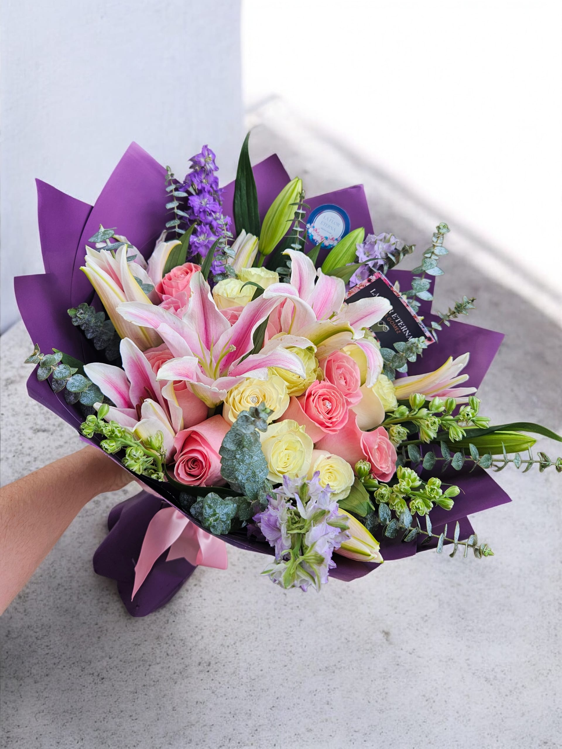Te Quiero - A bright, garden-fresh hand-tied bouquet that says "Te quiero" with every petal - perfect as a birthday bouquet, Mother's Day arrangement, anniversary roses alternative, or a cheerful congratulations gift. The mix of large pink lilies and clusters of pastel roses gives a romantic, uplifting impression the recipient will notice first: soft yellow and blush-pink roses soften the lilies' drama while spikes of purple delphinium add vertical movement and a playful pop.  This bouquet is gathered tightly for a layered, textured look and wrapped in bold purple paper, finished with a pale pink ribbon and gift tag for an elegant presentation. Small green buds and the delphinium spikes bring garden-like depth and contrast to the pale palette, making it feel both celebratory and intimate. Order to send someone a truly seen and celebrated moment - suitable for many occasions and ready to present straight from the shop.