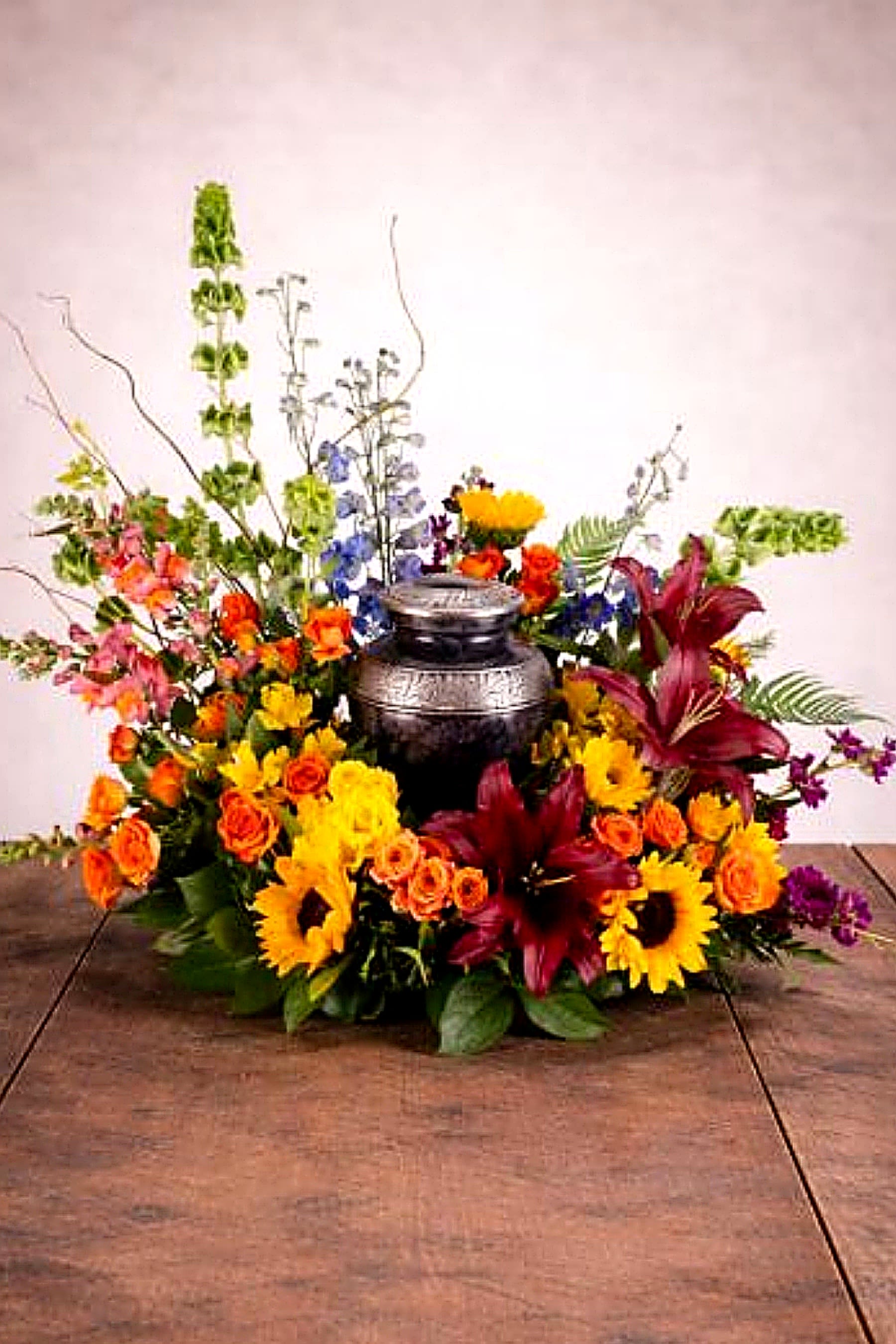 Dawn of Spring by BloomNation™ - The Dawn of Spring Arrangement from FloralFabulous is a radiant and uplifting cremation urn tribute designed to celebrate life, warmth, and new beginnings. This beautiful arrangement features a vibrant mix of sunflowers, roses, lilies, and colorful seasonal blooms, thoughtfully arranged around a central urn to create a meaningful and elegant presentation.  The bright and cheerful color palette symbolizes hope, renewal, and the celebration of a life well lived, making it an ideal choice for memorial services, celebrations of life, or honoring a loved one in a unique and personal way.  Designed to frame and complement a cremation urn, this arrangement creates a stunning focal point that brings comfort and light to family and friends. Its natural, garden-style design reflects beauty, warmth, and the lasting impact of cherished memories.  FloralFabulous proudly provides sympathy and cremation flower arrangements in Round Rock, TX and the greater Austin area, helping families express love, remembrance, and heartfelt support with care and compassion.