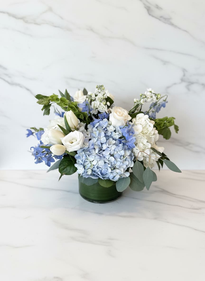 Low arrangement of white roses, tulips, and blue hydrangeas in a green glass cylinder vase