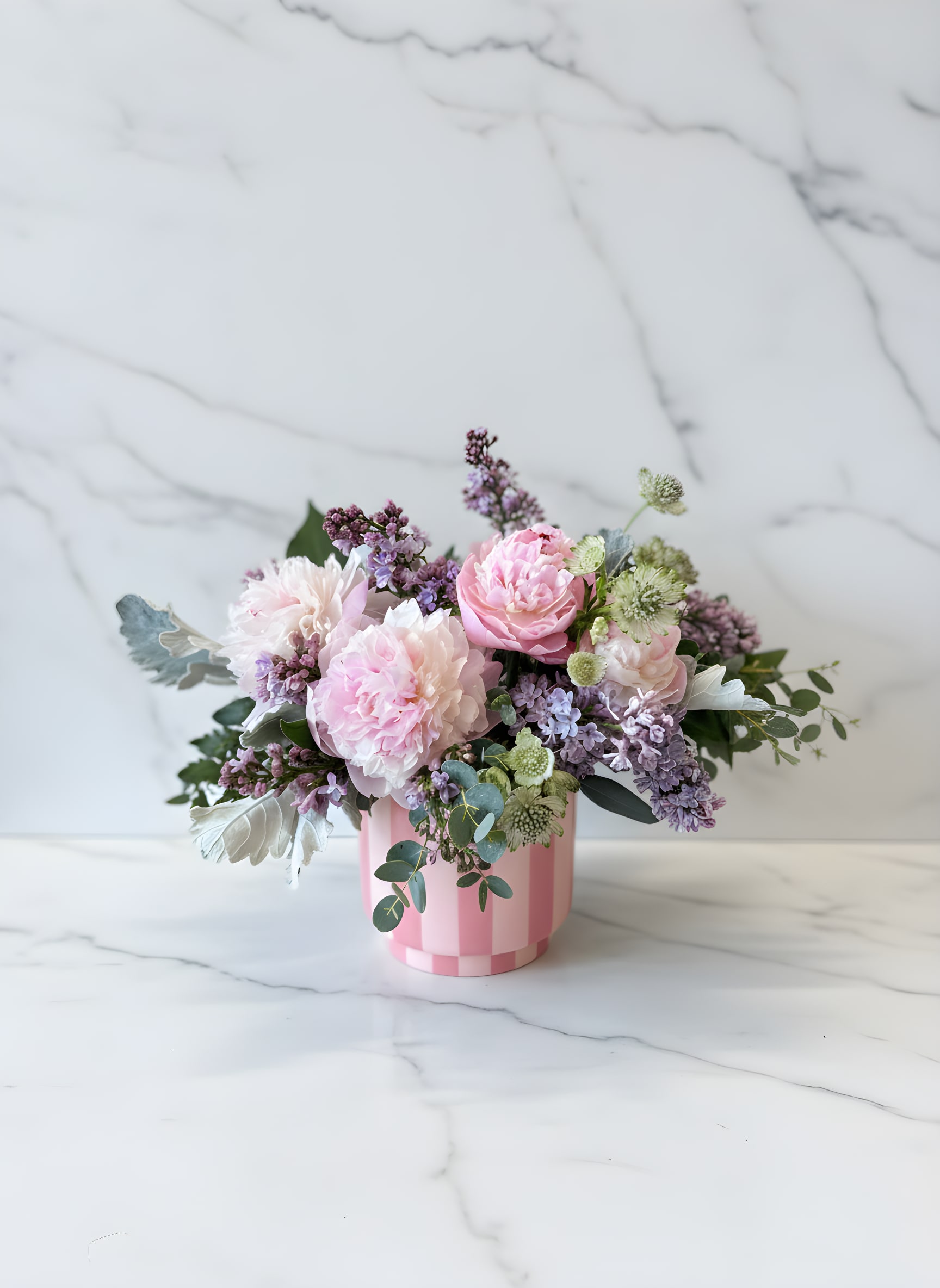 Petal Parfait  - A sweet and softly layered arrangement in a blush striped vessel, Petal Parfait feels like a spring desert for the eyes. Fluffy pink peony are nestled in a bed of fragrant lilacs and airy foliages to create a romantic collection with a playful twist.   Standard is approx. 12x12  Deluxe 15x15