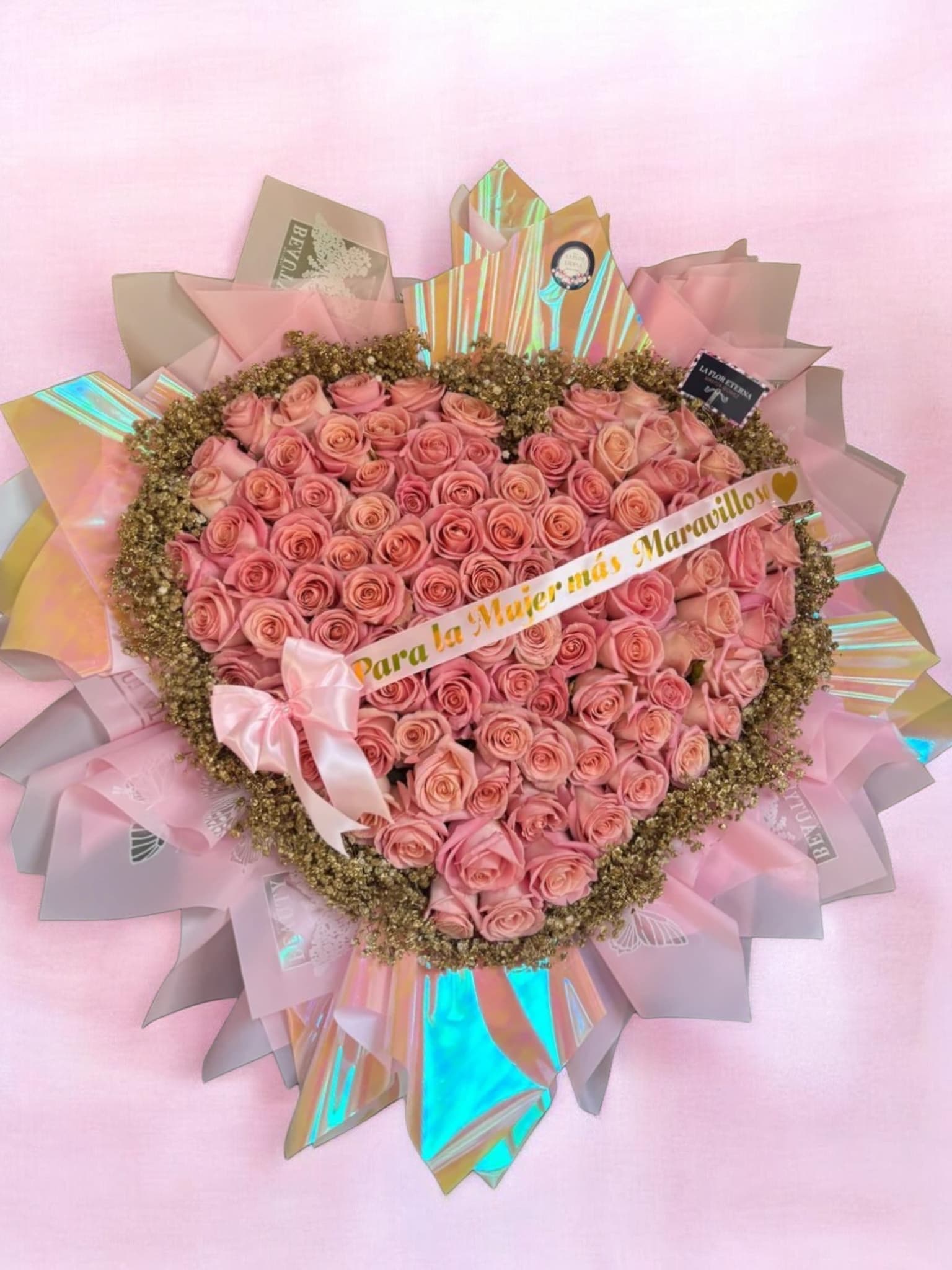 Pink Heart  - A lush heart-shaped arrangement of soft pink and peach roses forms a dense, romantic focal point-perfect as a birthday bouquet, an anniversary surprise, or a sentimental Mother's Day gift. The design reads like a declaration of affection for the woman you're honoring, the kind of arrangement that makes her feel truly seen and cherished for love and romance, "I'm sorry," or just because.  The heart is edged with a delicate ring of golden-toned filler flowers that add texture and vintage warmth while outlining the shape with subtle definition. Layers of iridescent and blush-pink wrapping paper fan out behind the blooms like celebratory rays, finished with a pink satin bow and a printed ribbon bearing a Spanish dedication for an extra personal touch. It arrives ready to present with an optional gift card-keep the roses fresh by changing the water daily and trimming stems for longest enjoyment.