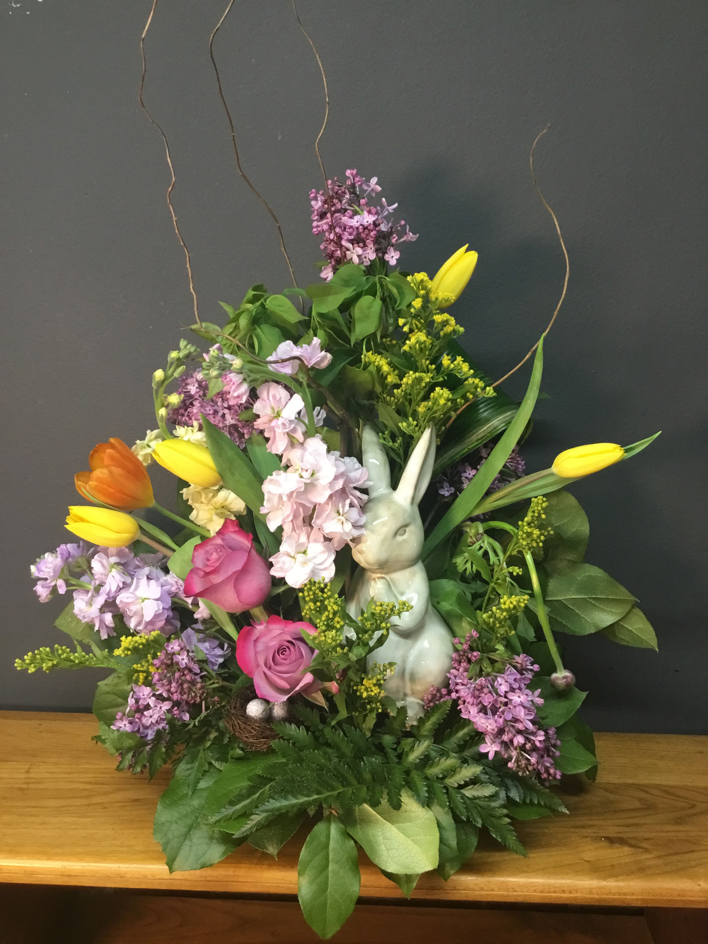 Spring Bunny Natural Arrangement - A white ceramic bunny tucked into a tall, fan-shaped spray of spring blooms makes this flower arrangement a joyful centerpiece for spring and an especially sweet Easter gift. The mixed-color palette-soft lilac clusters, blush pink roses, and vivid yellow and orange tulips-creates a playful, uplifting display that will make the person you're thinking of smile the moment it arrives.  This arrangement pairs fragrant stock and airy branches with classic tulips and roses for layered texture and scent. The bouquet's vertical silhouette gives it presence on a dining table or entry console; a decorative bird's nest with speckled eggs at the base reinforces the seasonal charm without feeling fussy. Sized to stand out but still easy to place, it's a bright, versatile choice for springtime décor or to celebrate a special someone who loves cheerful, garden-fresh style.
