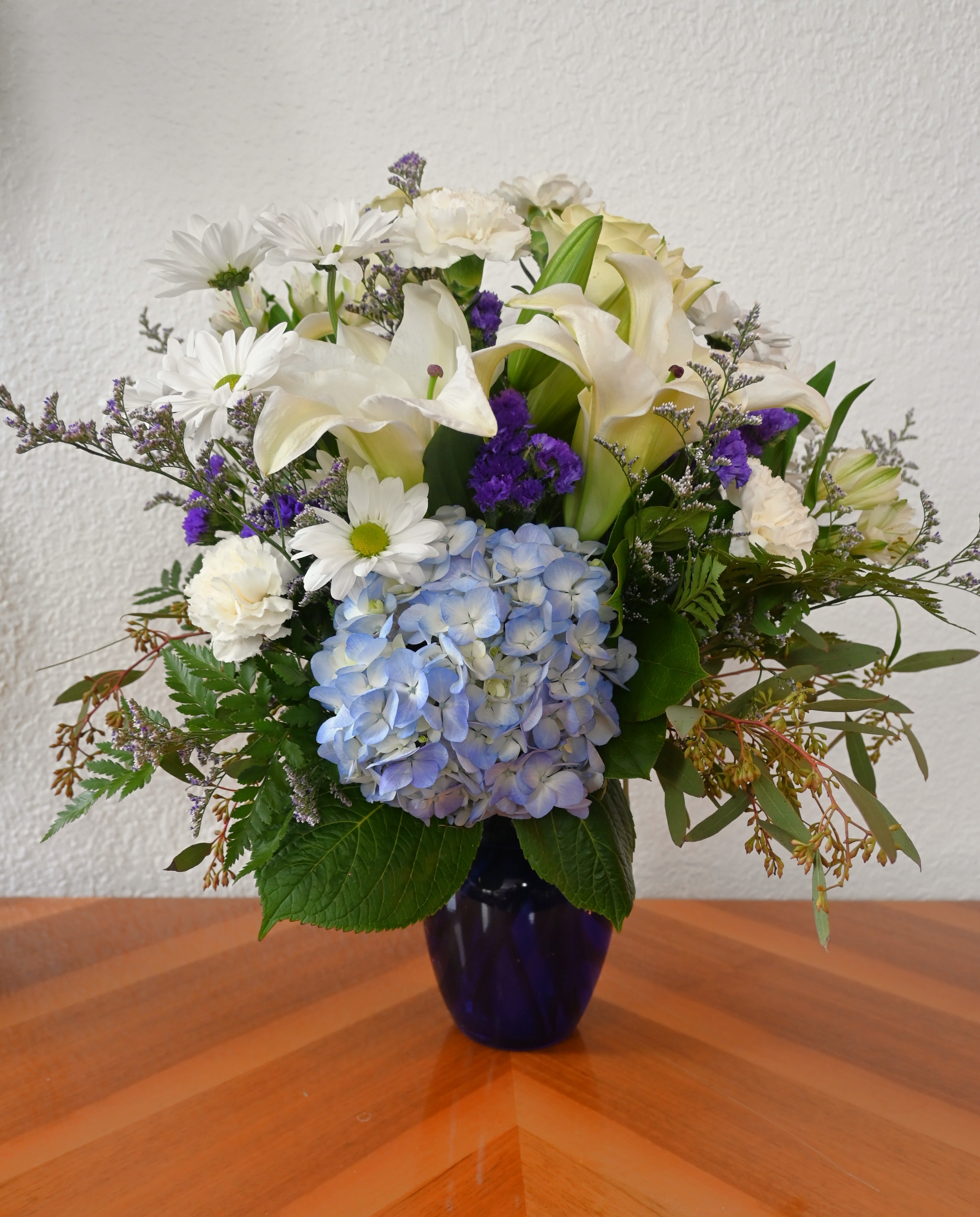 Blue Beauty - Brighten their home with the beauty of a bright blue sky. Arranged in a bold cobalt blue vase, this combination of blue hydrangea, white lilies, and more exudes peace and comfort.