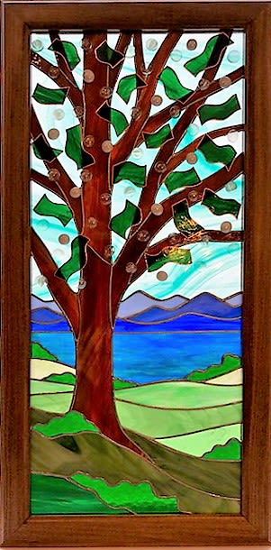 #A085 - Spring Day At The Lake - Stained Glass Window - A tranquil stained glass wall art piece that brings a lakeside spring morning into the home - perfect as a housewarming gift or a thoughtful everyday present for someone who loves nature-inspired décor. This wall-hung arrangement-like panel reads like a peaceful landscape: a tall brown tree with branching limbs and stylized green leaves stands against a bright, watery blue sky while rolling green hills and a deep blue lake stretch toward layered purple mountains. The calm palette of blues, greens, white and subtle accent tones creates a soothing focal point that will help the person you're thinking of feel comforted and at ease.  Crafted as a stained glass panel in a handmade poplar frame, the artwork includes circular glass accents tucked among the branches that catch and scatter light for gentle sparkle throughout the day. Sized to hang as wall décor, it's designed to fit living rooms, entryways, or cozy reading nooks where its reflective blues and layered scenery can brighten a room without overwhelming it. Choose this piece when you want to celebrate a new home, mark a quiet milestone, or give a gift that invites daily calm and reflection.