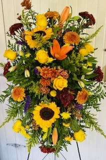 Sunflowers and lilies - Golden sunflowers and warm yellow roses form the first impression of this standing spray, designed for sympathy occasions or autumnal displays. The tall flower arrangement reads as a formal yet comforting tribute-ideal for memorial services, fall celebrations, or as a prominent statement in an entry or reception area-so the person you're honoring will feel remembered and seen.  This arrangement is densely filled with large sunflowers, yellow roses, orange lilies, and rust-colored chrysanthemums, with small purple accent blooms adding a gentle contrast to the golden and orange palette. Mounted on a metal easel stand, the spray's height and full texture create a bold but tender presence; the mix of broad sunflower faces, layered rose petals, and spiky lily accents gives both visual weight and delicate detail.  Appropriate when you want a respectful, seasonal expression of sympathy or a warm centerpiece for fall events, this standing spray arrives ready to display on its easel-no extra assembly required. To keep the flowers looking their best at a service or event, place the spray away from direct heat and check water foam if it will sit for extended periods.
