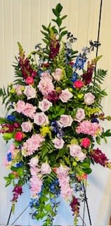 Wildflowers and Roses - A tall, elongated standing spray that reads as both graceful and consoling-this flower arrangement for sympathy and funeral services combines lavender roses with clustered pink blooms and deep burgundy snapdragons to create a layered, reverent presence. The inclusion of vivid blue delphinium spikes and airy stock adds vertical rhythm and color contrast, so the person you're honoring will be remembered with a composition that feels tender, thoughtful, and dignified.  Arranged on a metal easel, the design forms an oval silhouette that tapers at the top and bottom, with abundant greenery and trailing vines that cascade outward for a formal, dimensional effect. Colors range from mixed pastels and pinks to touches of green, blue, and red; the mix of roses, snapdragons, stock, and delphinium gives varied textures-velvety rose petals, tall snapdragon spires, and feathery delphinium-at a prominent standing height suitable for a chapel or graveside display.  Ideal for memorial services, wakes, or as a focal tribute at a funeral, this piece communicates sympathy and respect while drawing the eye with its calming palette and elegant structure. It arrives ready on its metal easel for immediate placement; to preserve blooms, keep the arrangement sheltered from direct sun and extreme heat until the service.