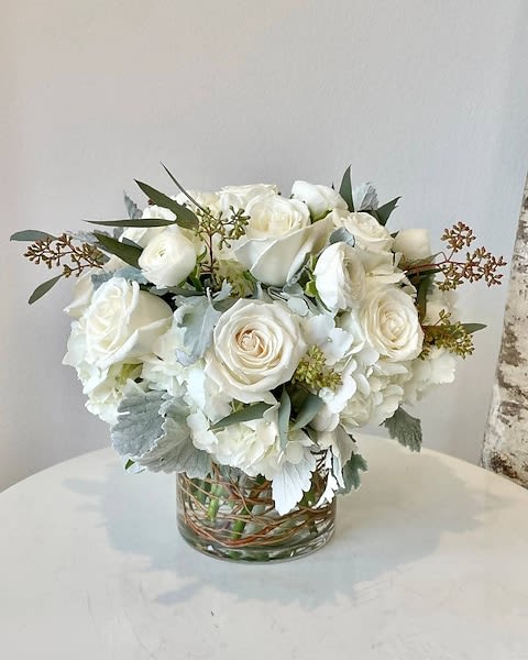 Angelic - A beautiful arrangement of ivory roses, white hydrangea, and white ranunculus sway gracefully in a timeless glass cylinder vase adorned with natural curly willow branches. The arrangement is further enhanced with frosty dusty miller and seeded eucalyptus greenery. The dimensions of this stunning 