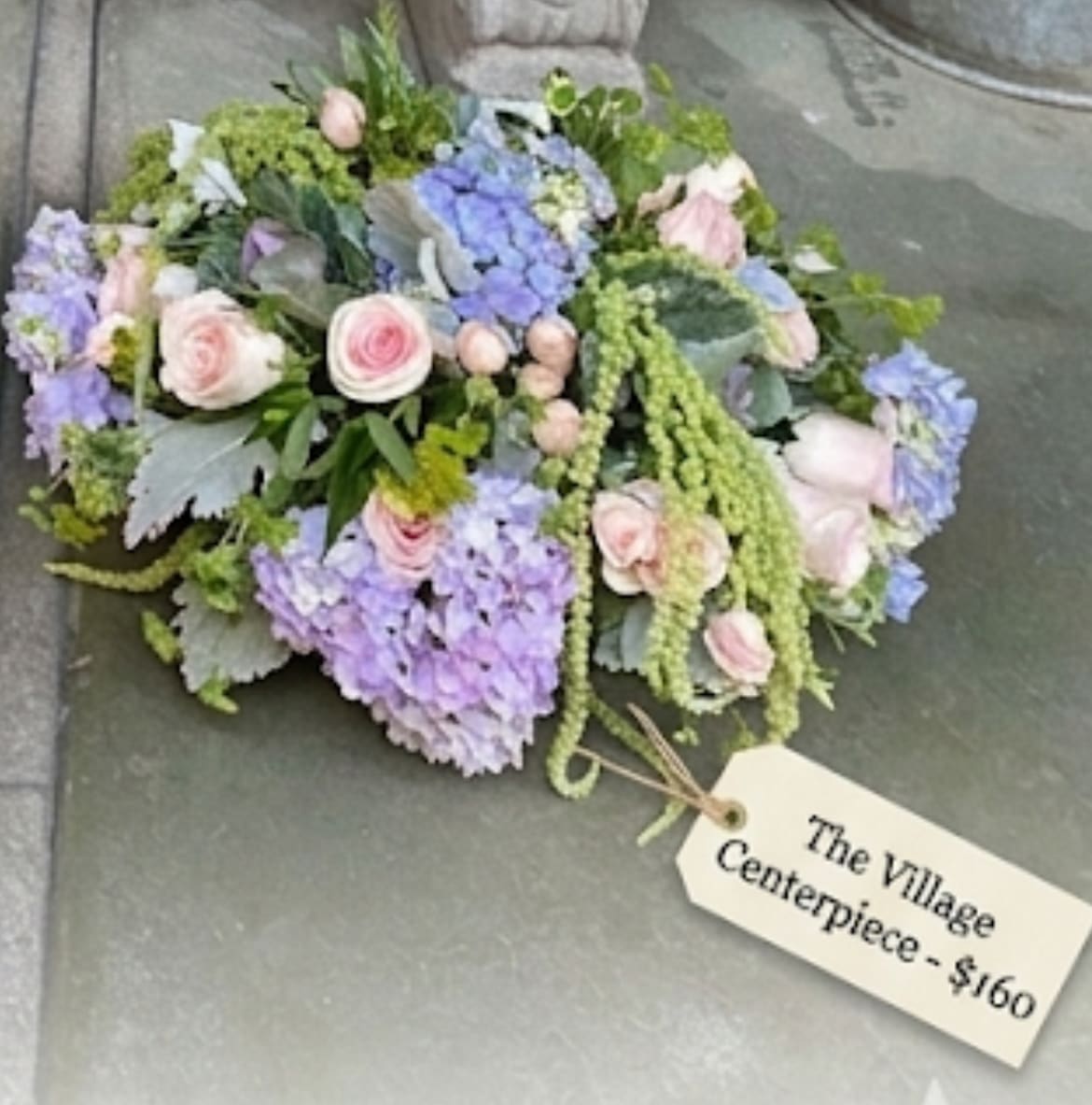 Village Centerpiece - A soft, garden-style arrangement of pale pink roses and clusters of blue and lavender hydrangea. Trailing greenery and textured foliage give it an airy, organic, and romantic feel.
