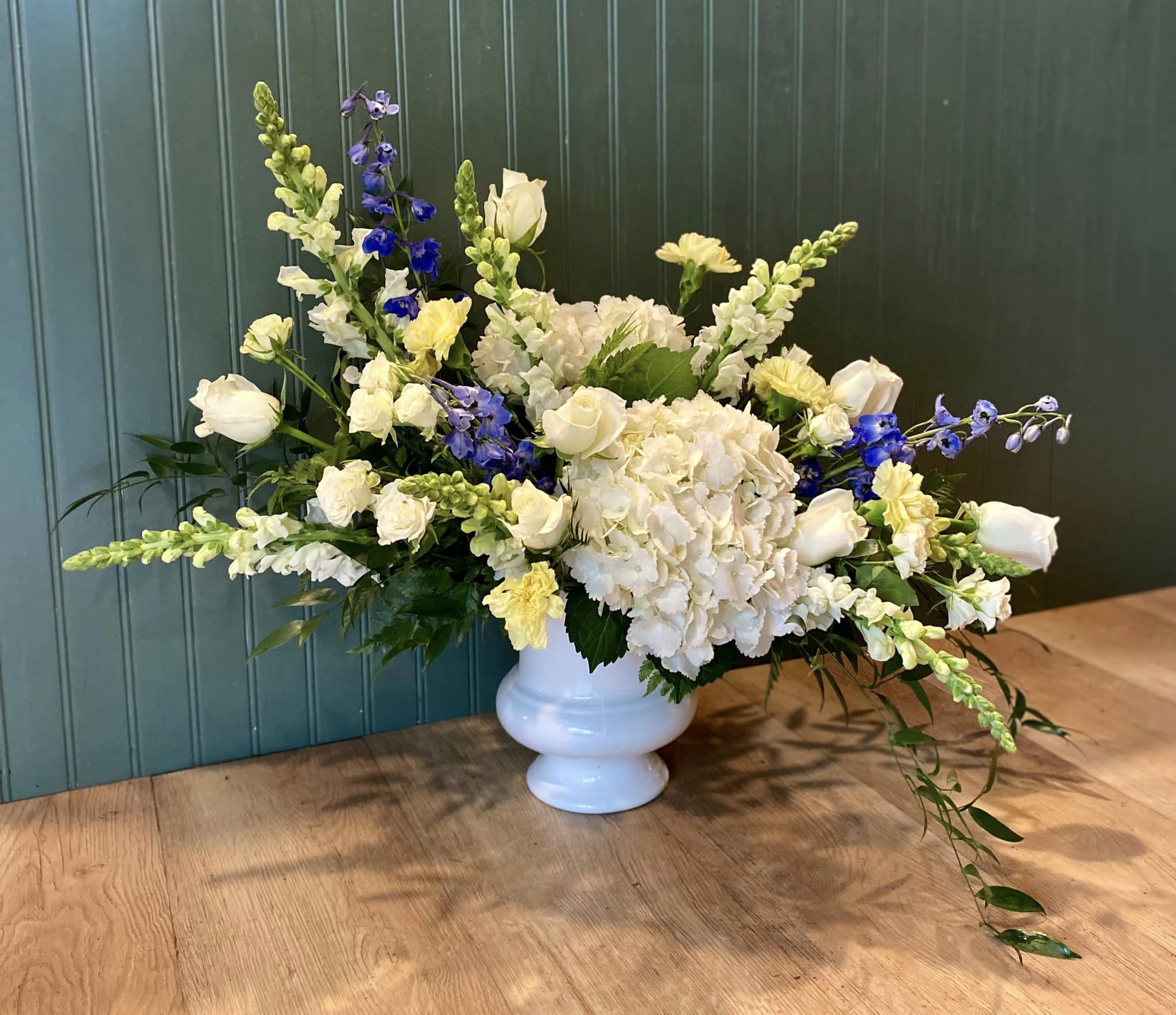 Wide white pedestal vase with white hydrangeas, roses, yellow blooms, and blue delphinium in an asymmetrical design