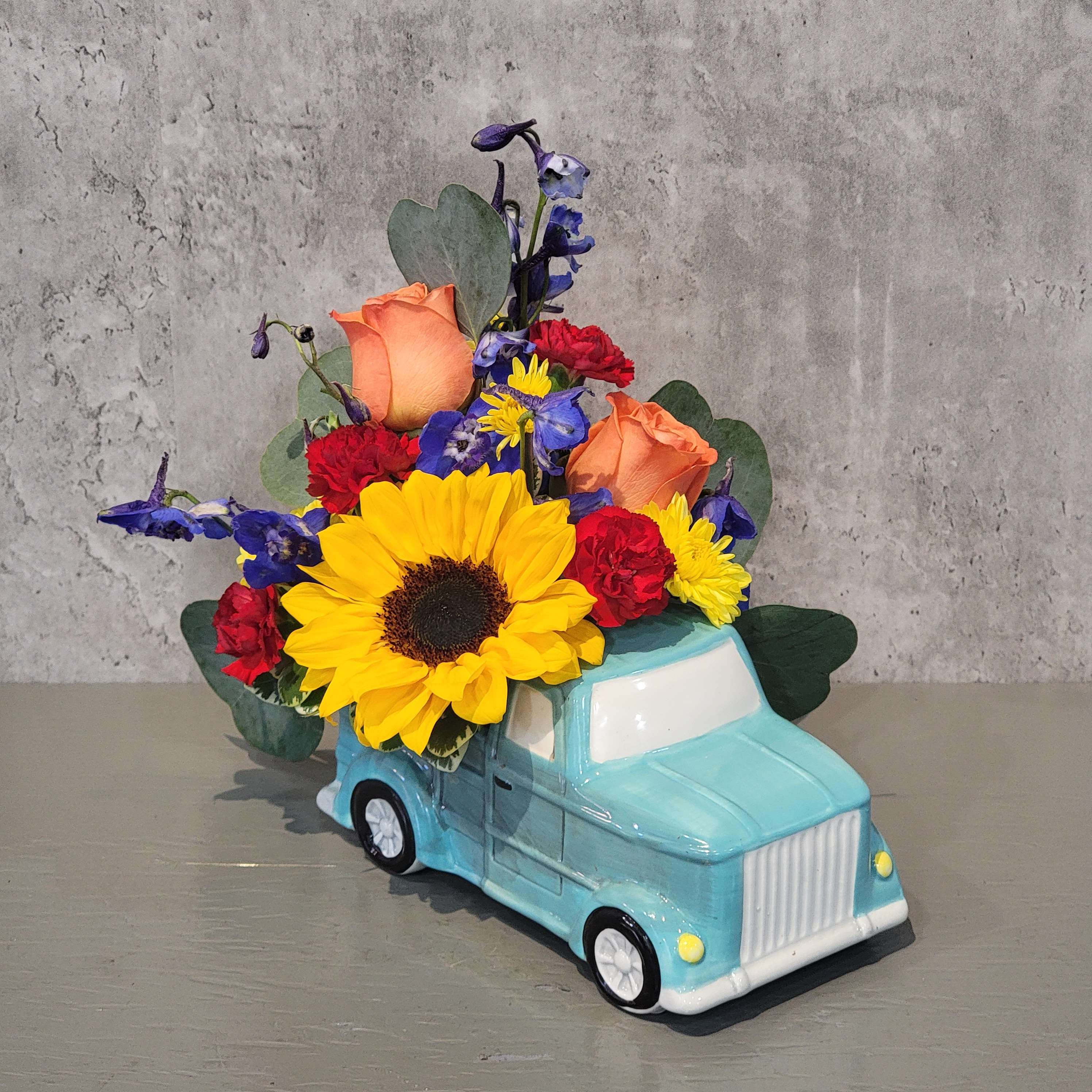 Trucking Along - Brighten their day and encourage them to keep 'trucking along' with this adorable ceramic turquoise blue truck, filled with eucalyptus, roses, sunflowers, carnations and mums. Perfect size to sit on the desk and remind your friend that you are thinking of them and believe in them! Arrangement measures approximately 8" tall and 9" wide. 