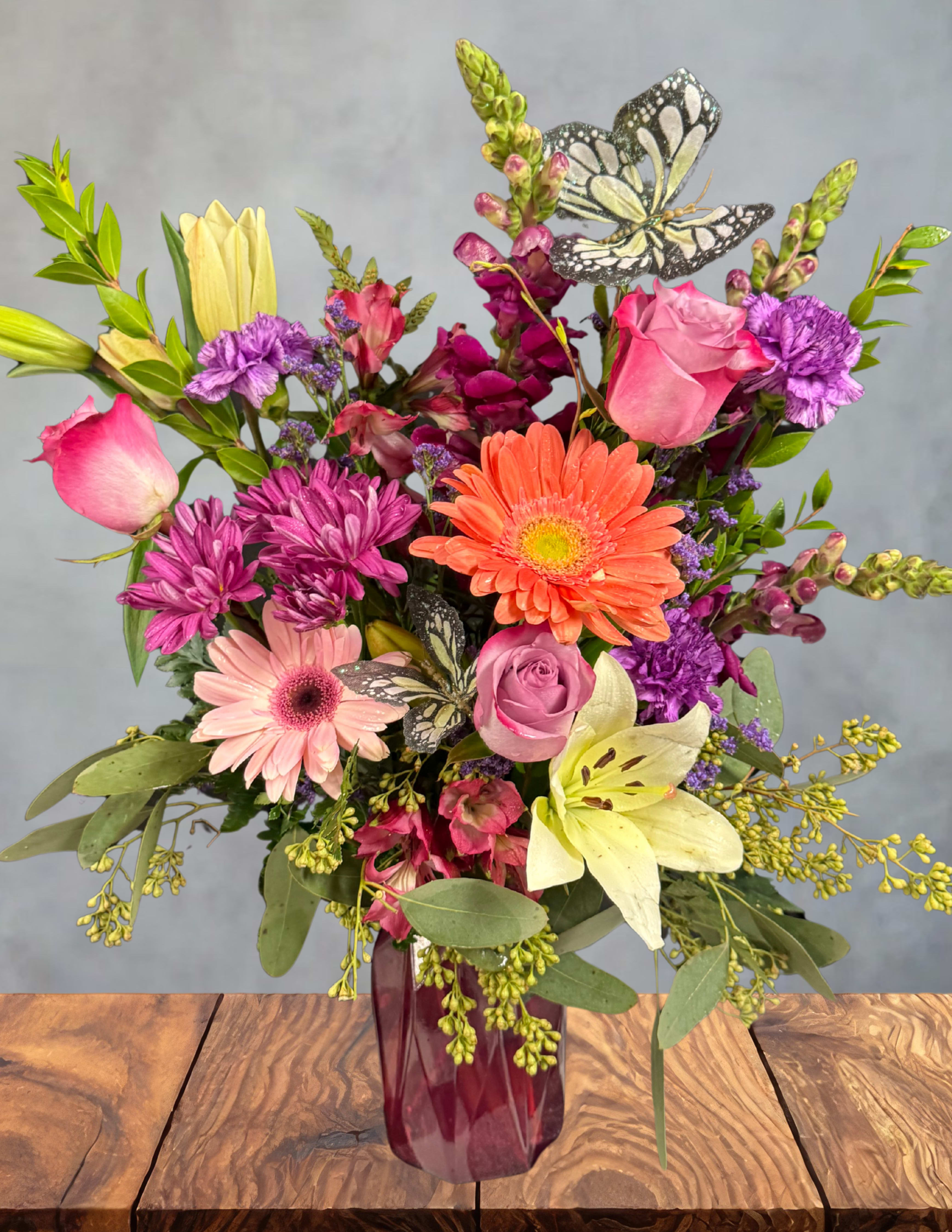 Pink Meadow -  Pink Meadow A cheerful burst of color inspired by a sun‑kissed garden in full bloom. Pink Meadow gathers vibrant roses, playful gerberas, lush carnations, and soft seasonal textures into a lively, uplifting display. Two delicate butterflies rest among the petals, adding a touch of whimsy and movement — as if they’ve just fluttered in to explore the blossoms.  Bright, joyful, and full of natural charm, this arrangement is perfect for celebrating happy moments, sending encouragement, or simply bringing a little meadow magic indoors.