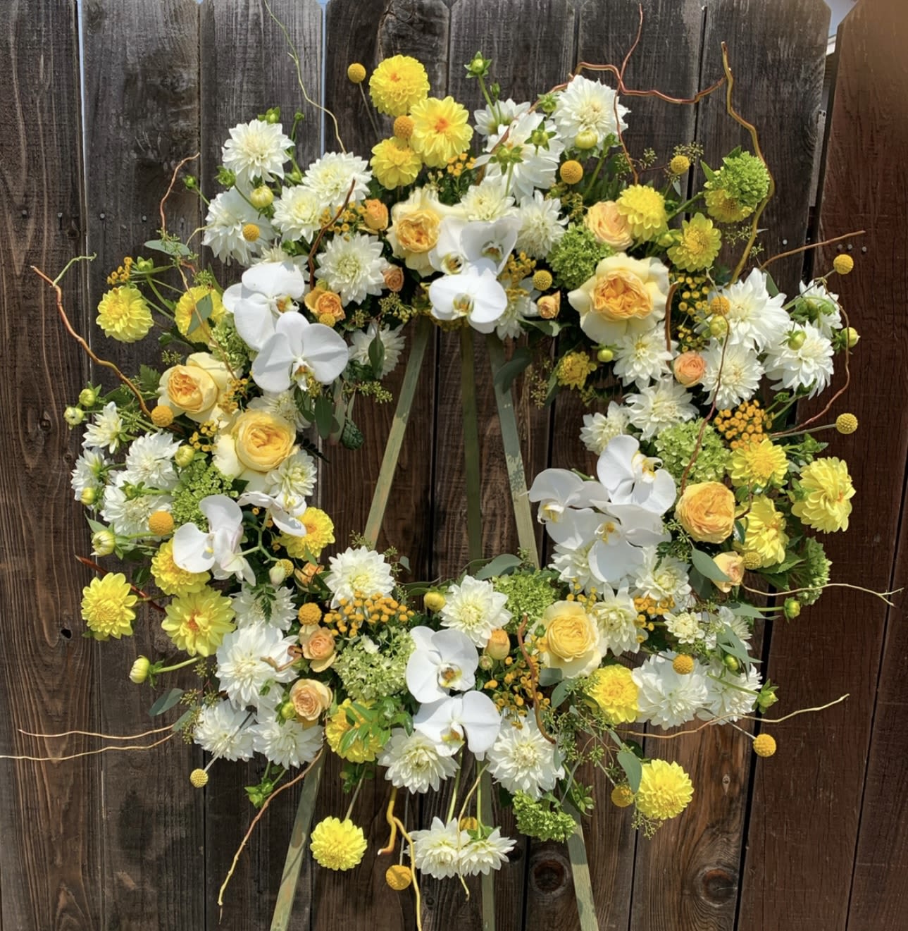 Dahlia Sunrise Wreath - Bright and uplifting, this standing wreath features a cheerful blend of yellow and white blooms including roses, orchids, dahlias, and craspedia. Accented with fresh greenery and natural curly willow, it's a radiant tribute perfect for honoring a joyful spirit and bringing comfort during times of remembrance.