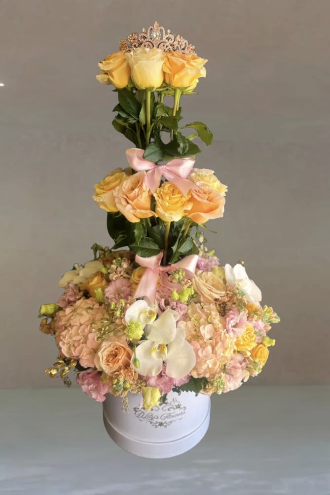 Crowned Blooms  by D'Lily's Flowers  - Where every bloom wears a crown A stunning three-tier arrangement of peach roses, soft pink hydrangeas, delicate orchids, and lush greenery, topped with a sparkling tiara and accented with pink satin bows. Elegant and regal, perfect for special occasions.