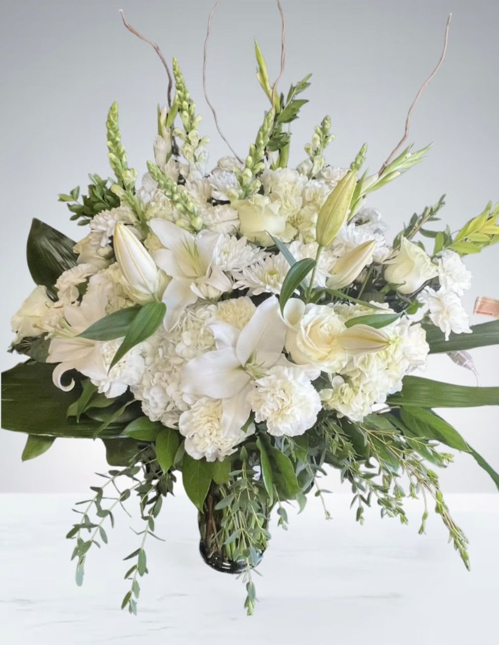 Serene Grace  by D'Lily's Flowers  - A timeless arrangement of white lilies, roses, snapdragons, and carnations, elegantly styled with lush greenery and curly willow branches. Perfect for sympathy, weddings, or peaceful home décor.