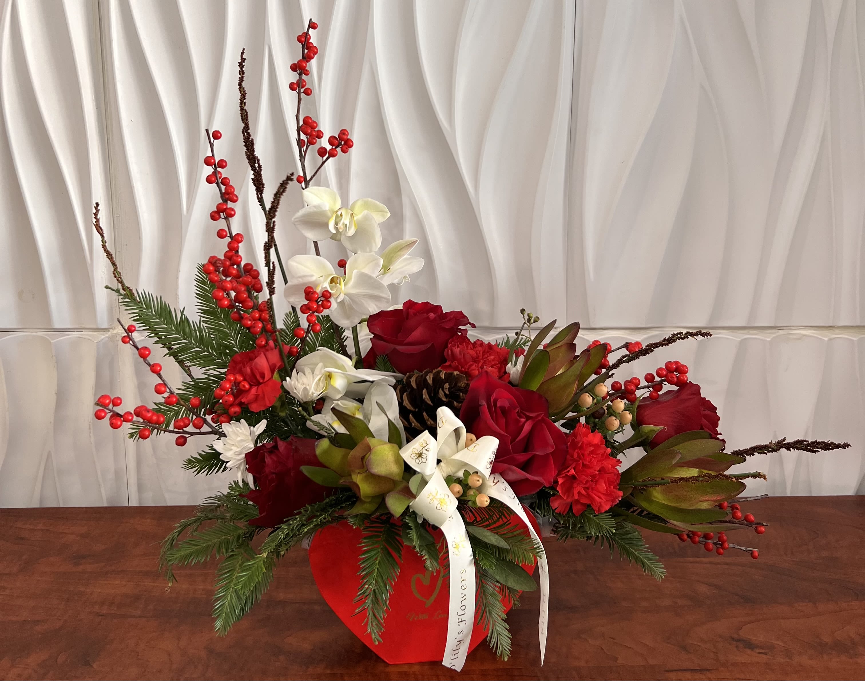 Christmas with Love - Designed in a festive red heart container, this Christmas arrangement celebrates love, warmth, and togetherness with red florals, white orchids, and lush seasonal greenery.