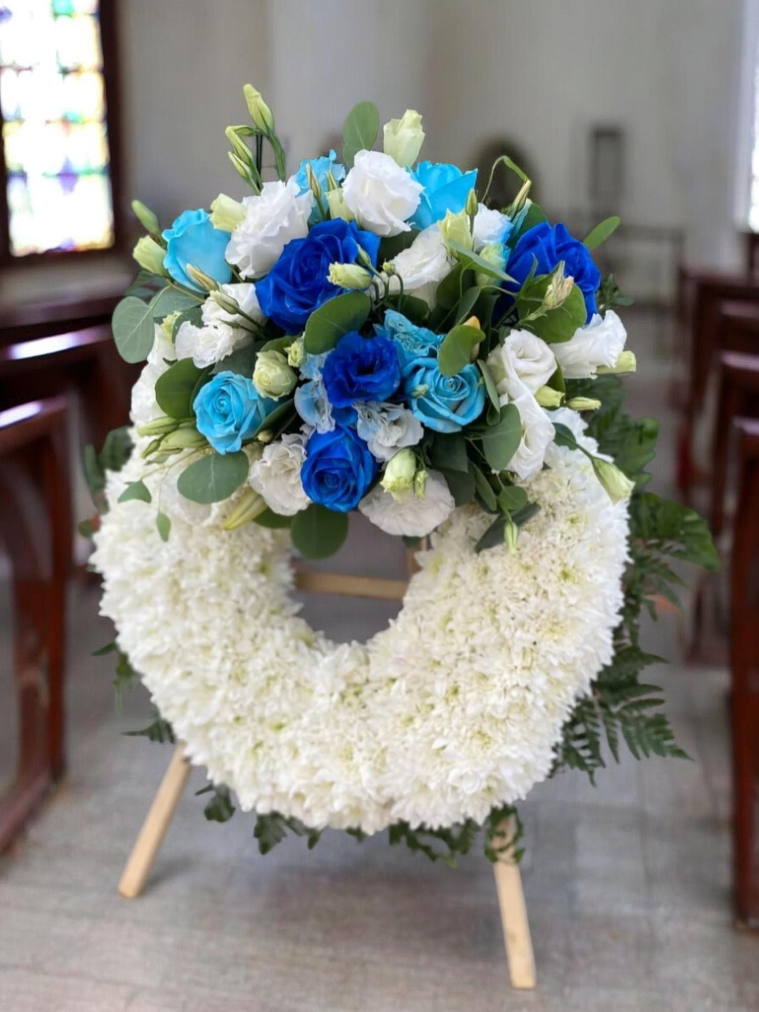 Peaceful Blue  - A full ring of densely packed white chrysanthemums forms a calm, formal standing funeral wreath designed to offer comfort at memorials and remembrance services. This flower arrangement for a funeral places the white chrysanthemum ring as the visual anchor, while a compact spray of vivid blue and white roses with soft white lisianthus at the top provides a gentle, consoling contrast-perfect for honoring a loved one with quiet dignity.  The wreath shows off crisp white chrysanthemums throughout, accented by blue roses, white roses, and delicate lisianthus buds and blooms clustered at the crown. Mounted on a simple wooden easel stand, the arrangement has the height and presence needed for chapel or service display; the cool blue, white, and green palette reads as respectful and serene. The circular form symbolizes continuity and remembrance, making this an appropriate tribute for funerals and memorial services.