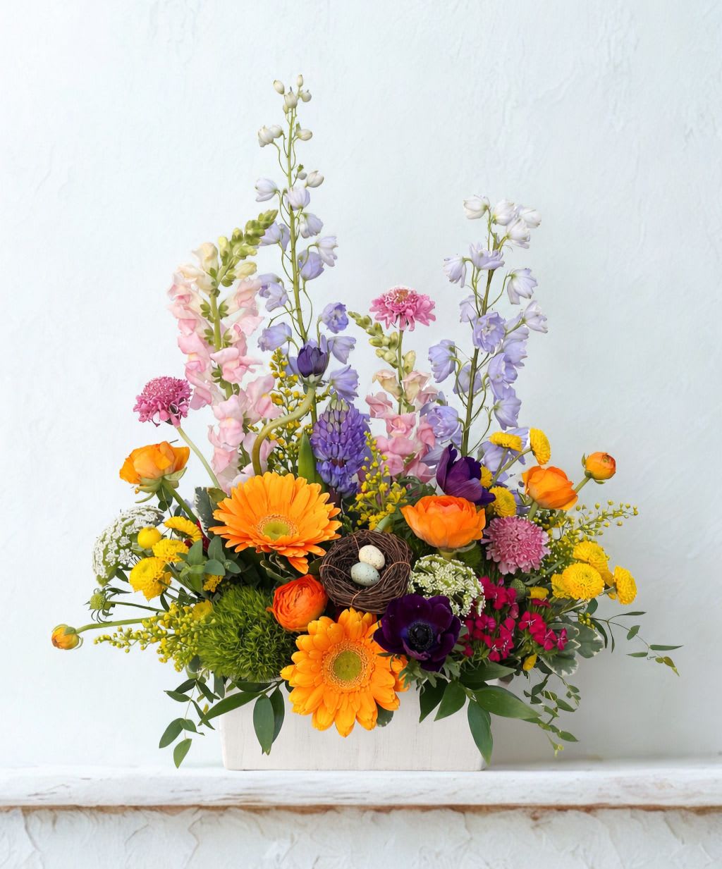 Colorful spring flower arrangement with orange gerberas and mixed blooms in a white box with a small bird nest