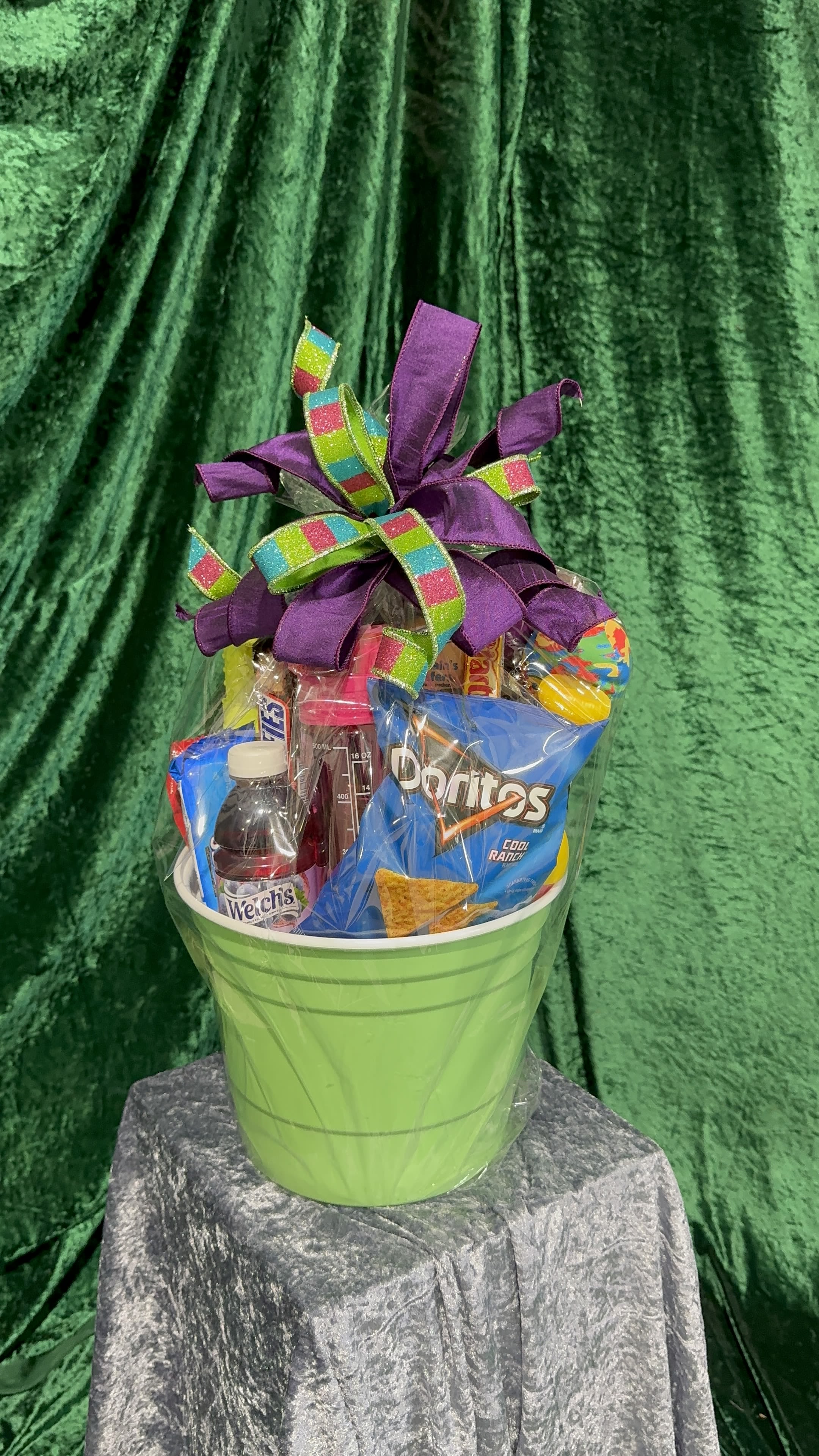 Easter Baskets - kids - Bright, playful and ready to delight, this kids' Easter basket is a casual gift-perfect for birthday surprises, get well wishes, or a cheerful holiday treat. The arrangement comes in a bright green plastic bucket and is wrapped in clear cellophane so the colorful snack bags and drinks peek through-she'll feel instantly celebrated when the multicolored goodies and large purple bow come into view.  Packed with assorted packaged snacks, candy, gum, and crackers plus a bottled juice drink and a clear pink sports bottle, the mix is lively and kid-friendly. A small toy ball on a stick adds a playful finishing touch, and the bow-deep purple ribbon with a glittery multicolor checkered accent-gives the whole presentation an extra festive pop. The bucket measures as a handheld gift-size that's easy for kids to carry and enjoy right away.  Perfect for an Easter basket alternative, classroom party favor, or a bright "thinking of you" surprise, this ready-made set is designed to spark smiles with minimal fuss. Because it's pre-wrapped in cellophane, it's ready to give as soon as you receive it-no additional assembly required.