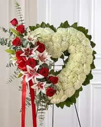 ALWAYS IN MY HEART - A standing heart-shaped funeral wreath that speaks of enduring love and gentle remembrance, designed for memorial and sympathy services. The heart form and red-and-white palette make this sympathy arrangement an unmistakable expression of deep affection and honor for the person you're remembering.  Densely covered in small white chrysanthemums for a soft, textured background, the arrangement is accented on one side by a cluster of rich red roses and white lilies with red centers. A red satin bow with long trailing ribbons hangs from the floral cluster, giving the piece a formal, commemorative feel. The wreath is mounted on a metal easel stand for a tall, prominent presence that reads well at the front of a service or beside a casket.  Made to convey love and comfort to mourners, this design is an appropriate tribute when you want to say "always in my heart." The standing size and easel make delivery and display straightforward; if you need assistance with delivery timing or placement at the service, our shop can coordinate those details.