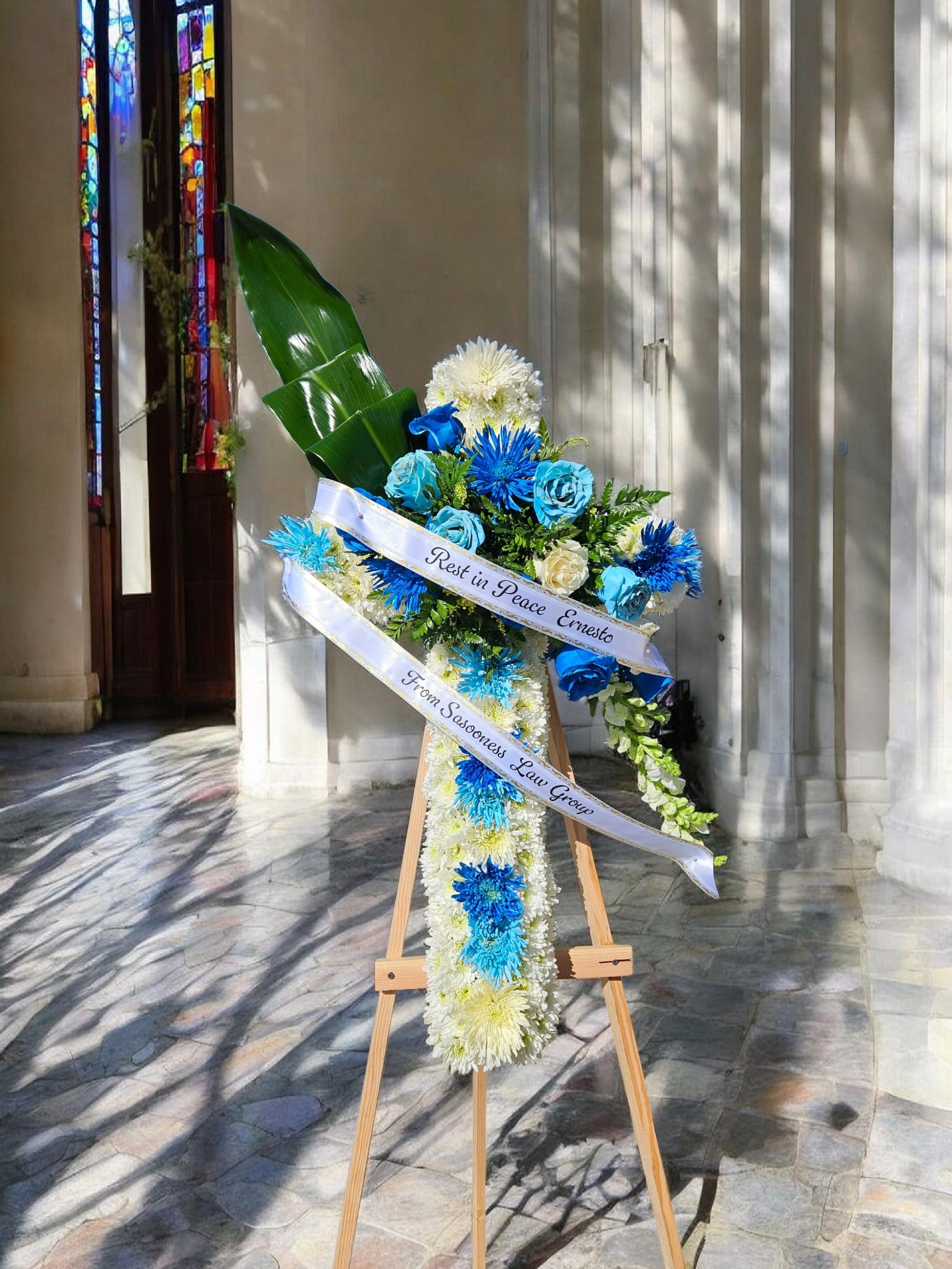 Cruz de Madera  - A tall wooden easel holds a dignified sympathy cross covered in a dense carpet of white chrysanthemums with vertical bands of blue blooms down the center - a funeral standing arrangement designed to convey reverence and comfort at memorial services. The printed white ribbon carries a respectful message, making this tribute piece an immediate signal of condolence for a service, graveside farewell, or church memorial.  Clusters of blue roses and white flowers gather at the upper right for visual emphasis, lifted by a large glossy green leaf that adds height and contrast against the chrysanthemum backdrop. The palette of white, blue, yellow, and green reads classic and calming; the cross's proportions are tall and upright on its simple wooden easel to ensure visibility in a sanctuary or reception area.  Made to honor and console, this arrangement is intended for those who want to express sympathy with solemn beauty - the kind of tribute that helps the bereaved feel seen and supported. The printed ribbon is applied as shown; please specify any personalization or message at checkout so the memorial text appears exactly as requested.