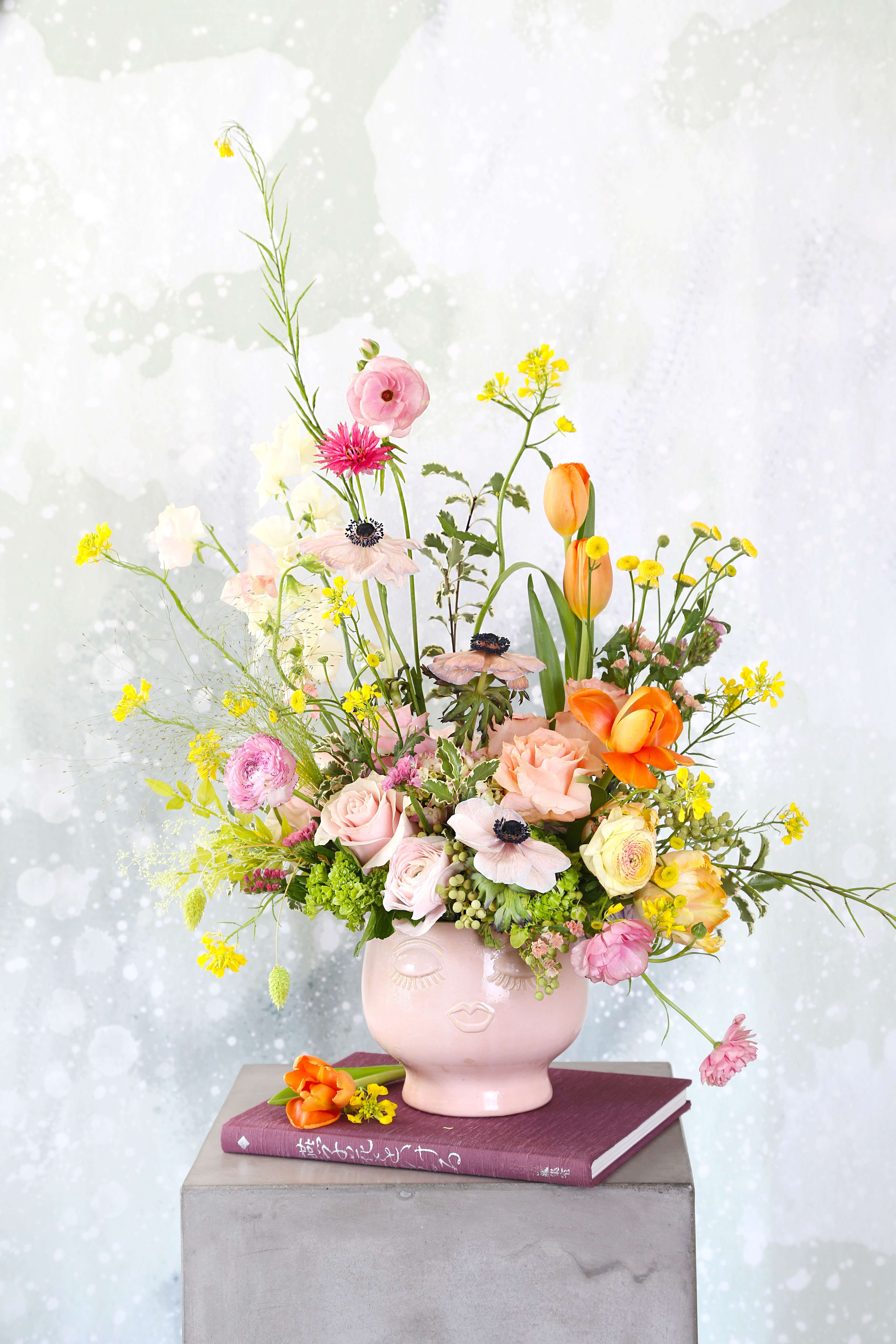 Mommy-Limited Edition - Brighten your space with our enchanting Whimsical Floral arrangement, featuring a delightful mix of seasonal blooms. Nestled in a charming blush face-shaped ceramic vase., this arrangement showcases soft pink garden roses, vibrant orange tulips, delicate anemones, Dutch Hydrangea, and cheerful yellow wildflowers, all artfully combined to create a stunning display. This arrangement will deliver with gift box and greeting card.  