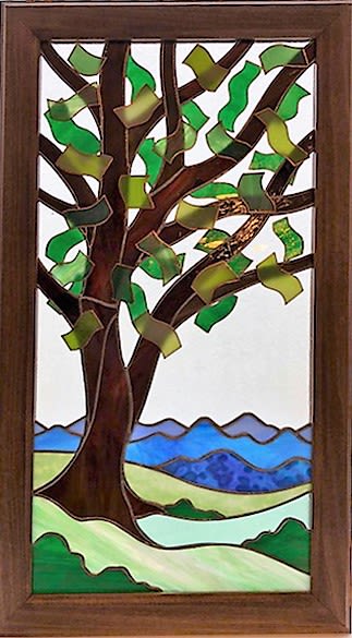 #A102 - Summertime Twin Set (left twin) - Stained Glass Window - A vertical stained-glass panel that reads like a quiet summer morning - this left twin in the Summertime Twin Set is the mirror image of it’s twin, making them perfect to hang in sidelight windows.  This framed stained glass art piece is perfect as a heartfelt Mother's Day gift, a thoughtful housewarming present, or a calming addition for someone you want to encourage. The stylized tree and rolling hills create an immediate sense of peace, so the person you're thinking of will feel seen and comforted every time light plays across the glass. Handcrafted within a wood frame, the panel uses curved brown glass for the trunk and branches and ribbon-like green and yellow-green pieces to suggest fluttering leaves.   The lower scene layers translucent greens for hills, a pale river, and distant blue-purple mountains; the mix of opaque and translucent glass catches daylight differently throughout the day, adding depth and a contemplative glow. Sized to display vertically on a wall or window, it pairs naturally with occasions from anniversaries and birthdays to retirement, sympathy, "I'm sorry," or simple "thinking of you" moments. This piece works year-round-give it for Father's Day, Christmas, a wedding, or "just because" to mark new beginnings-and it especially suits anyone who appreciates quiet, nature-inspired décor. The wood-framed panel arrives ready to hang; no special care is required beyond occasional dusting to keep the glass bright and the scene luminous.