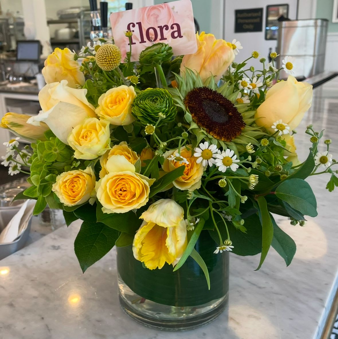 Mellow Yellow - Roses, sunflowers, ranunculus, spray roses, feverfew and tulips in shades of yellow and green in a vase.