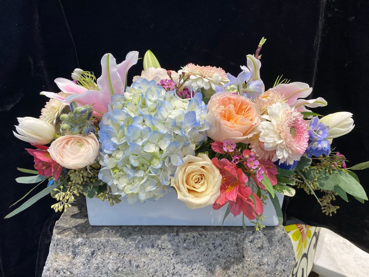 For Your Easter Table - A simple white bisque ceramic rectangle makes a home for a selection of Spring blooms for the Easter table. Some of the favorites this time of year include blue hydrangea, pink mini Gerbera daisies, Premium roses and a selection of textural greenery are sure to make an elegant centerpiece. Celebrate the holiday with Crimson Petal's creations!