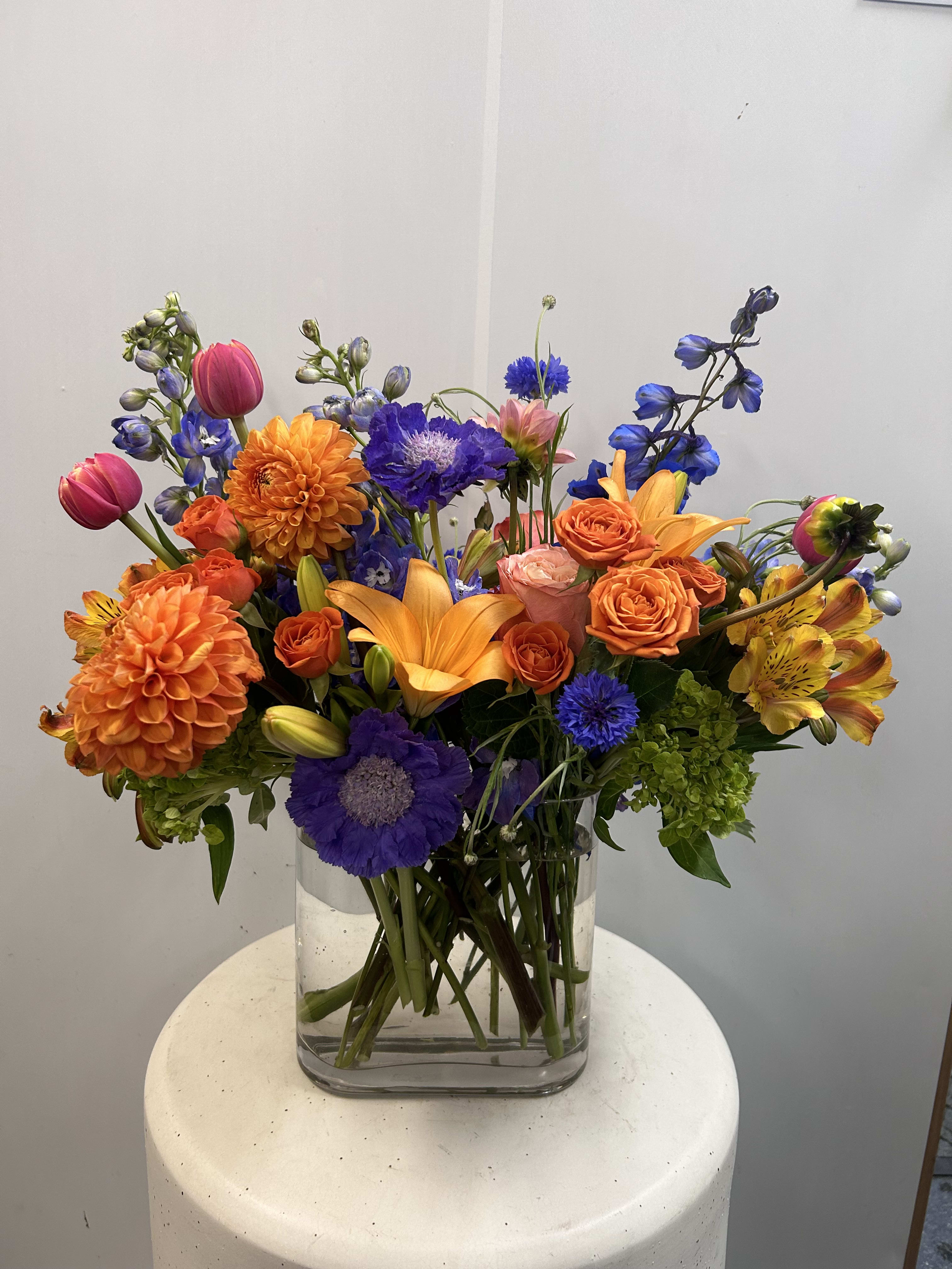Miles Apart - A beautiful complimentary combination of bold oranges and blue hues. Created in a mid height glass with a mixture of seasonal flowers. 