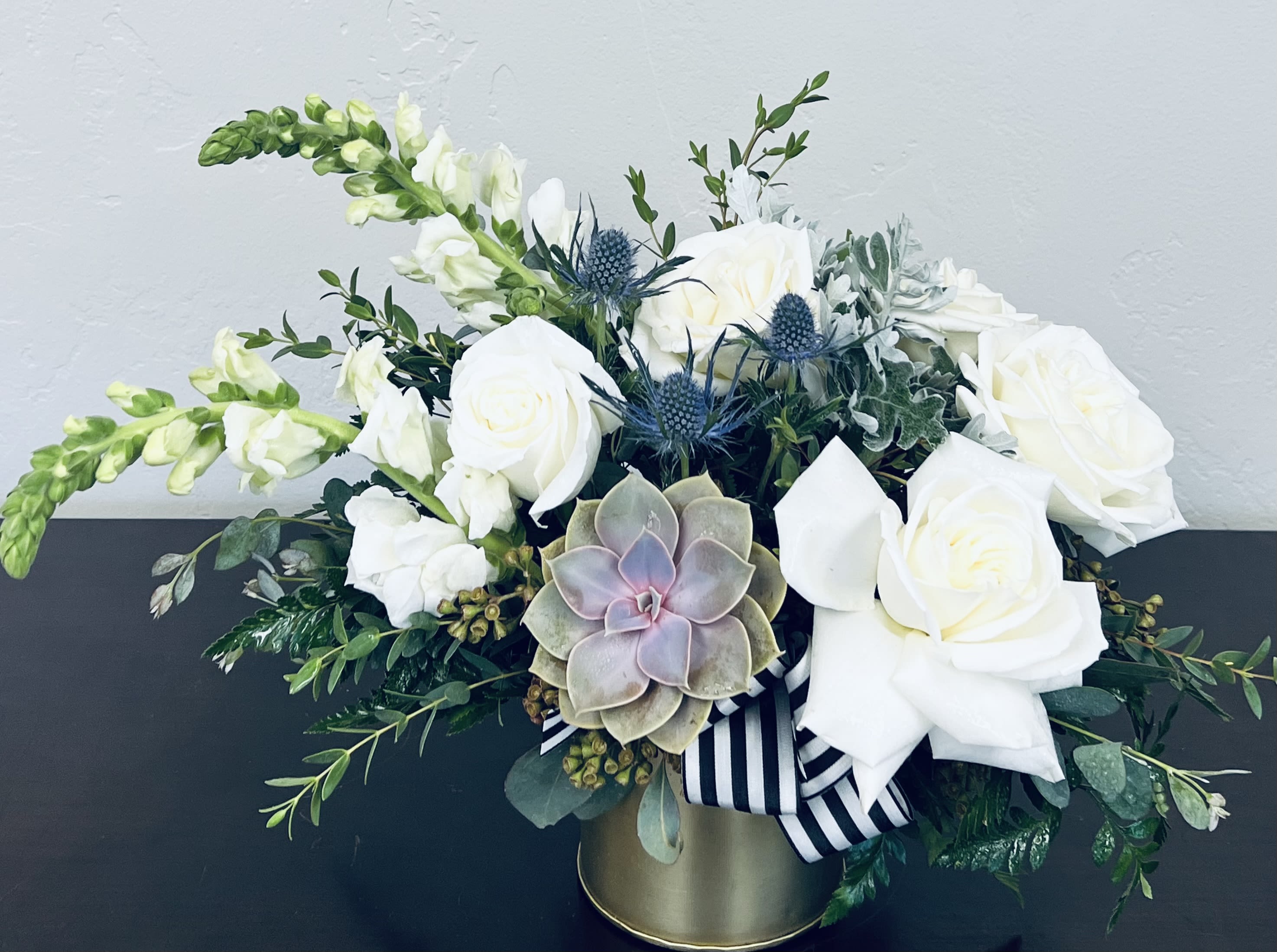 Mountain Bloom -  The Mountain Bloom   Soft, romantic, and effortlessly elegant—this design overflows with creamy whites, silvery textures, and lush greenery. Nestled within is a succulent you can keep and grow forever, making this piece as lasting as it is beautiful. Bigger than ever, we have added 2 additional roses and more foliages to make this arrangement more luxurious.  Arranged in a vintage-inspired vessel and finished with a striped ribbon, the Mountain Blossom Arrangement brings together rustic charm and feminine grace. A lush floral that feels both timeless and unforgettable. 