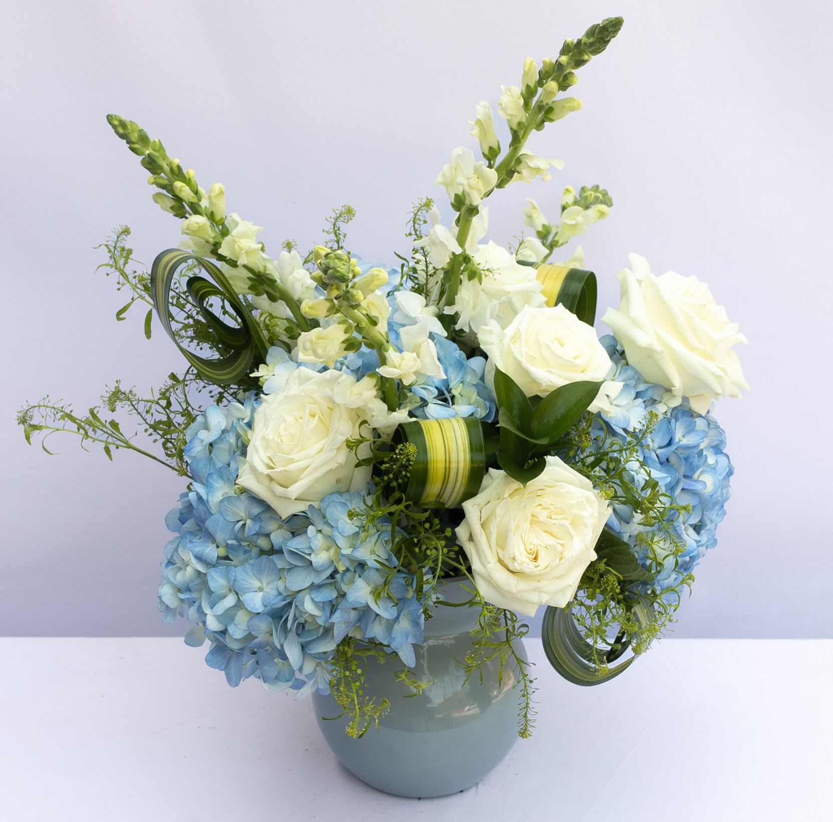 The Luxurious Bouquet - Clusters of blue and green hydrangea give this floral arrangement a fresh focus drawing the eye with pops of white roses and snapdragons perfectly placed with an artist’s eye to create an unforgettable moment upon delivery. Notice how the flax leaves are placed within the arrangement to give it all a sense of movement and grace taking the design aesthetic to a new level of sophistication. The subtle light blue color of the vase helps to bring forth the blues and greens within the bouquet making this both a warm and airy design that will help you send a gift that truly speaks from your heart. 