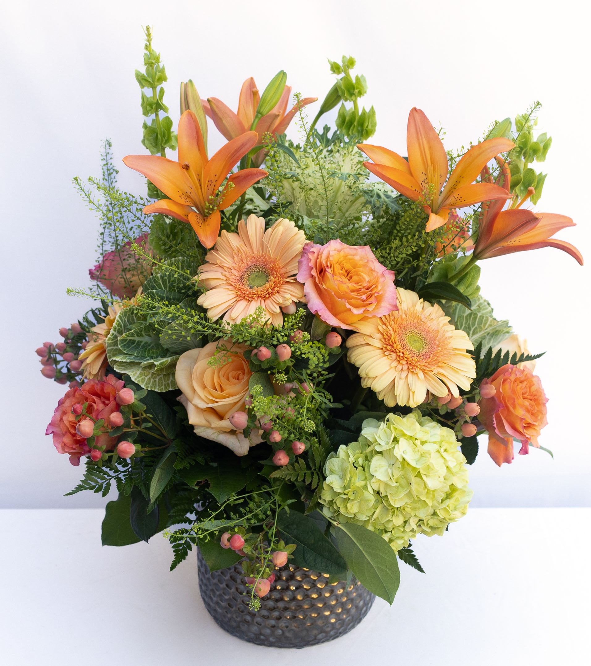 Peachy Keen  - Shades of Pink, Peaches and Pistachio Greens arranged in a garden-style assortment of hydrangeas, bells of Ireland, roses, cremons, lilies, greenery, fillers, &amp; berries. Arranged in a cooper mercury vase that is 9hx7w.  The arrangement designed is about 25hx20w.   