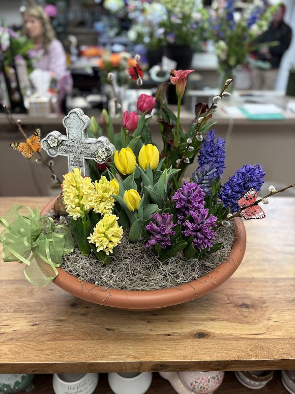 Blooming Bulb Garden with Cross - A cheerful low bulb garden arranged in a rustic terracotta bowl-this Blooming Bulb Garden with Cross blends purple, yellow, pink, and fresh green for an uplifting Easter centerpiece or a gentle sympathy gift. Clusters of vivid purple and pale yellow hyacinths mingle with bright yellow and pink tulips, while a single pink calla lily adds a graceful vertical note. The engraved stone-like cross pick rises from one side to give quiet meaning, and tiny butterfly picks flutter through the moss for a whimsical, hopeful touch.  Planted in a wide terracotta bowl and nestled in soft moss, the design is finished with a green sheer ribbon bow at the rim to reinforce the fresh spring palette. Dimensions are low and wide to sit nicely on a table or mantel; the bulbs are arranged for immediate color and ongoing bloom as the bulbs settle. This is a thoughtful way to say "thinking of you" at Easter, for a memorial service, or simply to bring warm, living color into someone's home.  Haehn Florist, Greenhouses, &amp; Flower Delivery stands behind every substitution: if a specific bulb variety is unavailable we'll replace it with blooms of similar color, texture, and seasonal quality to preserve the arrangement's look and meaning. Order now for delivery or pickup from Haehn Florist at 410 Hamilton Rd. Wapakoneta. Follow us on Facebook: www.facebook.com/haehnflorist