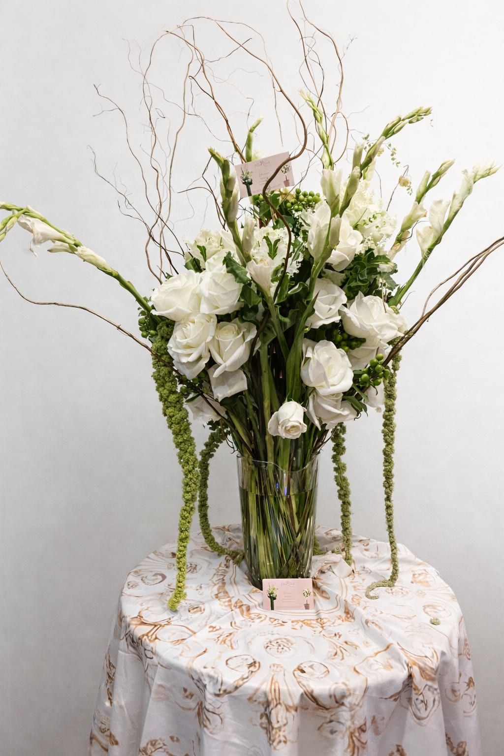 Tall white flower arrangement with roses and trailing green accents in a clear glass vase on a round table.