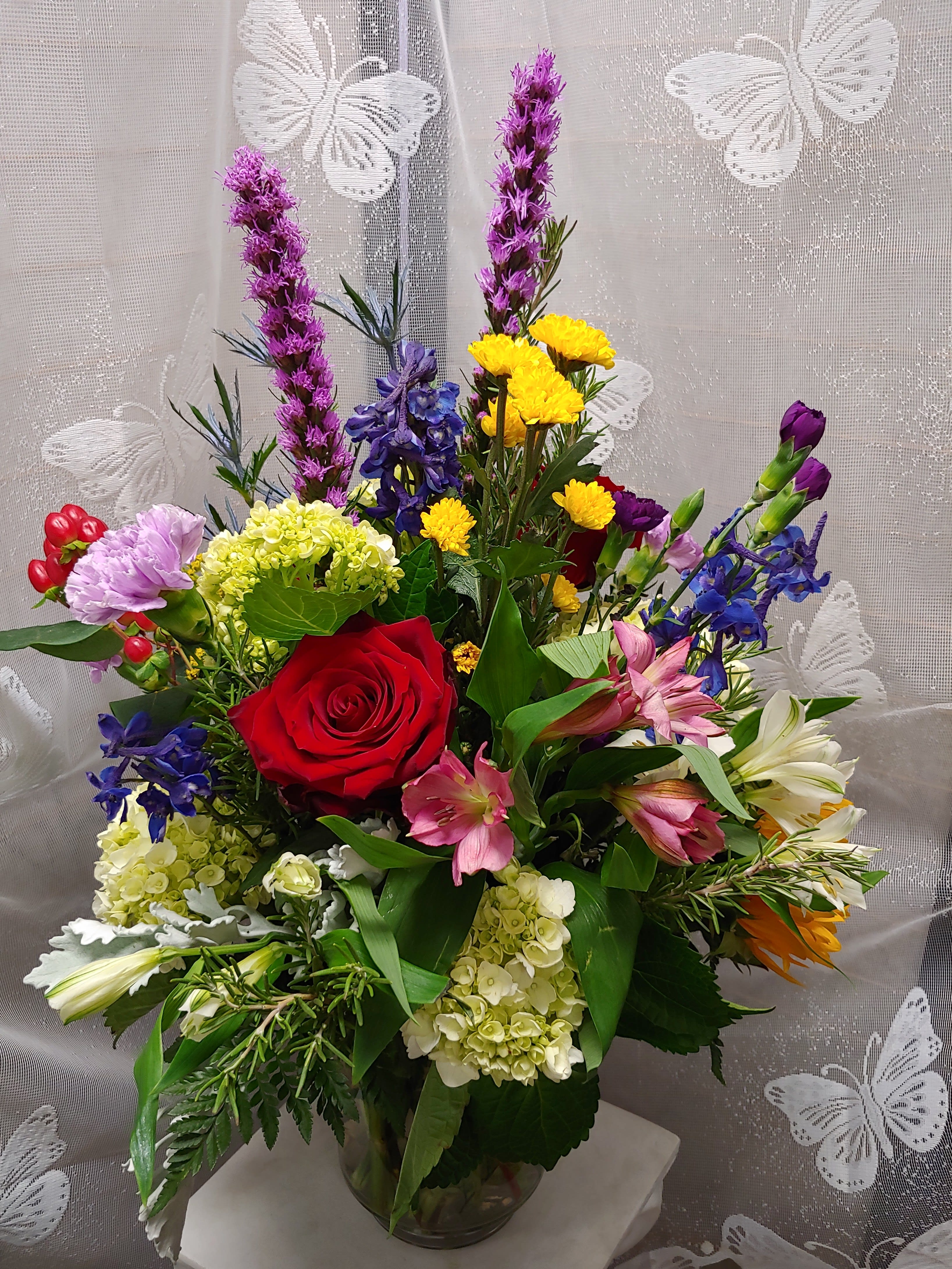 Garden party - A tall, colorful Garden Party flower arrangement that brings lively, celebratory energy to birthdays, anniversaries, or a thoughtful "just because" surprise. A bold red rose anchors the center, surrounded by pink alstroemeria and pale hydrangea clusters that give the bouquet a soft, romantic heart-perfect for love and romance or a meaningful anniversary gift. The mix also reads comforting and sincere for sympathy, get well, or an "I'm sorry" gesture, so the person you're thinking of will feel genuinely seen and uplifted.  This mixed arrangement features red roses, white lilies, pink alstroemeria, lavender carnation-like blooms, purple flower spikes and blue delphinium for vertical drama, plus small yellow chrysanthemums and daisies that brighten the middle. Arranged in a clear glass vase, the stems create a tall, garden-fresh silhouette that looks effortless on a dining table or bedside stand. The varied colors-red, purple, blue, yellow, and mixed tones-make it versatile for Thank You bouquets, Best Friends Day or Grandparent's Day surprises, and even Boss's Day or Valentine's Day.  Easy to display and enjoy, the Garden Party is designed to feel cheerful and welcoming in any season. For longer enjoyment, change the water every few days and trim stems at an angle when refreshing the vase; available in a single standard size that fits most tabletops and entryway consoles.
