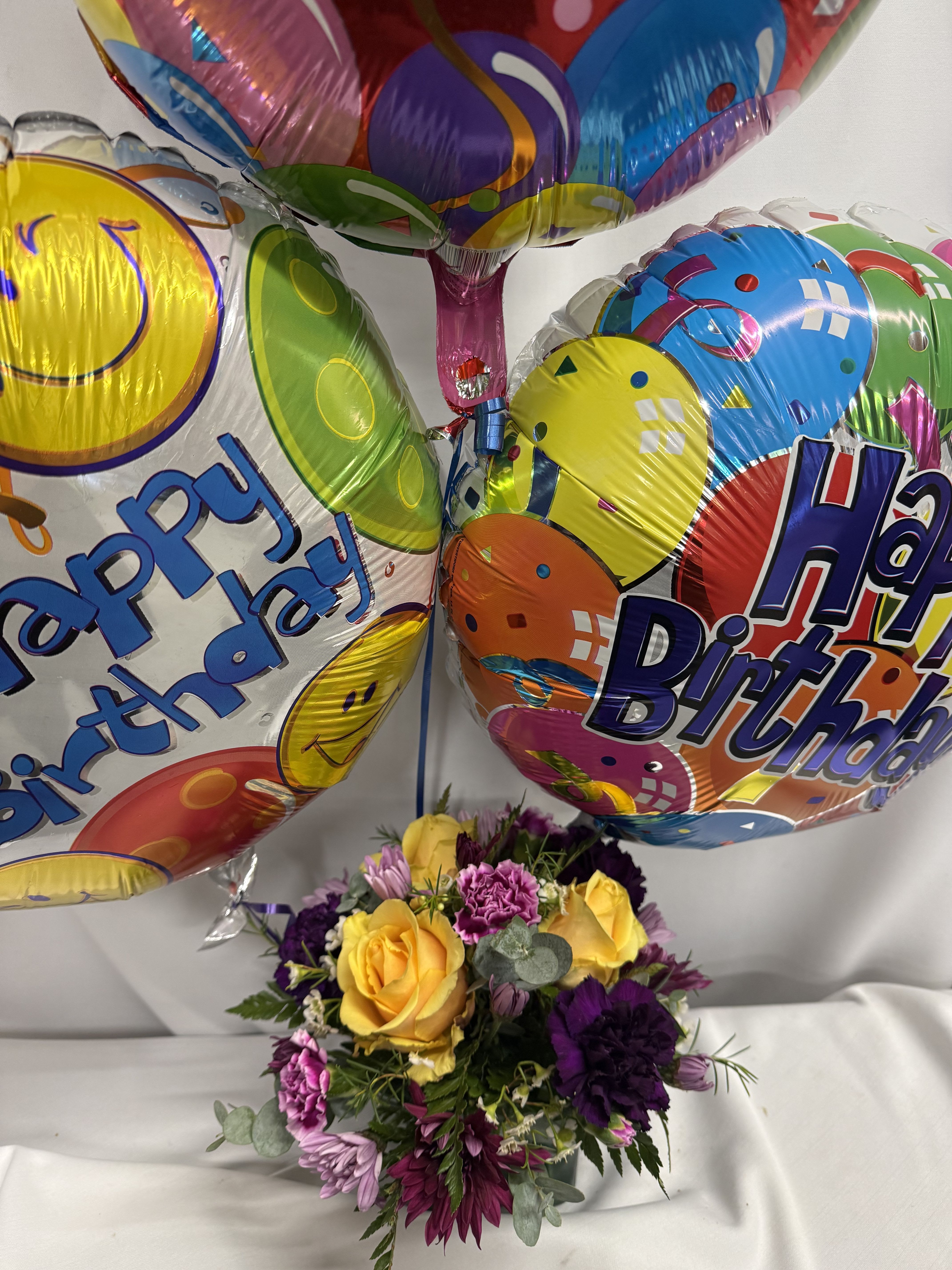 It’s your Birthday!  - Bright, cheerful yellow roses set the tone for this birthday flower arrangement, paired with purple and pink alstroemeria, carnations, daisies and chrysanthemums to create a playful, high-contrast bouquet perfect for a birthday celebration. The compact mixed bouquet arrives in a clear 4 x 4 glass cube, topped with three large round "Happy Birthday" mylar balloons-colorful patterns and smiley faces rising above the blooms to make the recipient feel celebrated the moment they walk in the room.  This arrangement mixes soft textures and bright hues-velvety rose petals, frilly carnations, and daisy-like mums-with long-lasting alstroemeria to keep the display lively for days. The cube vase gives it a modern, countertop-ready footprint that's ideal for desks, dining tables, or bedside stands. It's a joyful way to say "happy birthday" or "congratulations" to a friend, family member, or coworker; select delivery timing at checkout and enjoy simple care: keep water fresh and out of direct midday sun to extend bloom life.