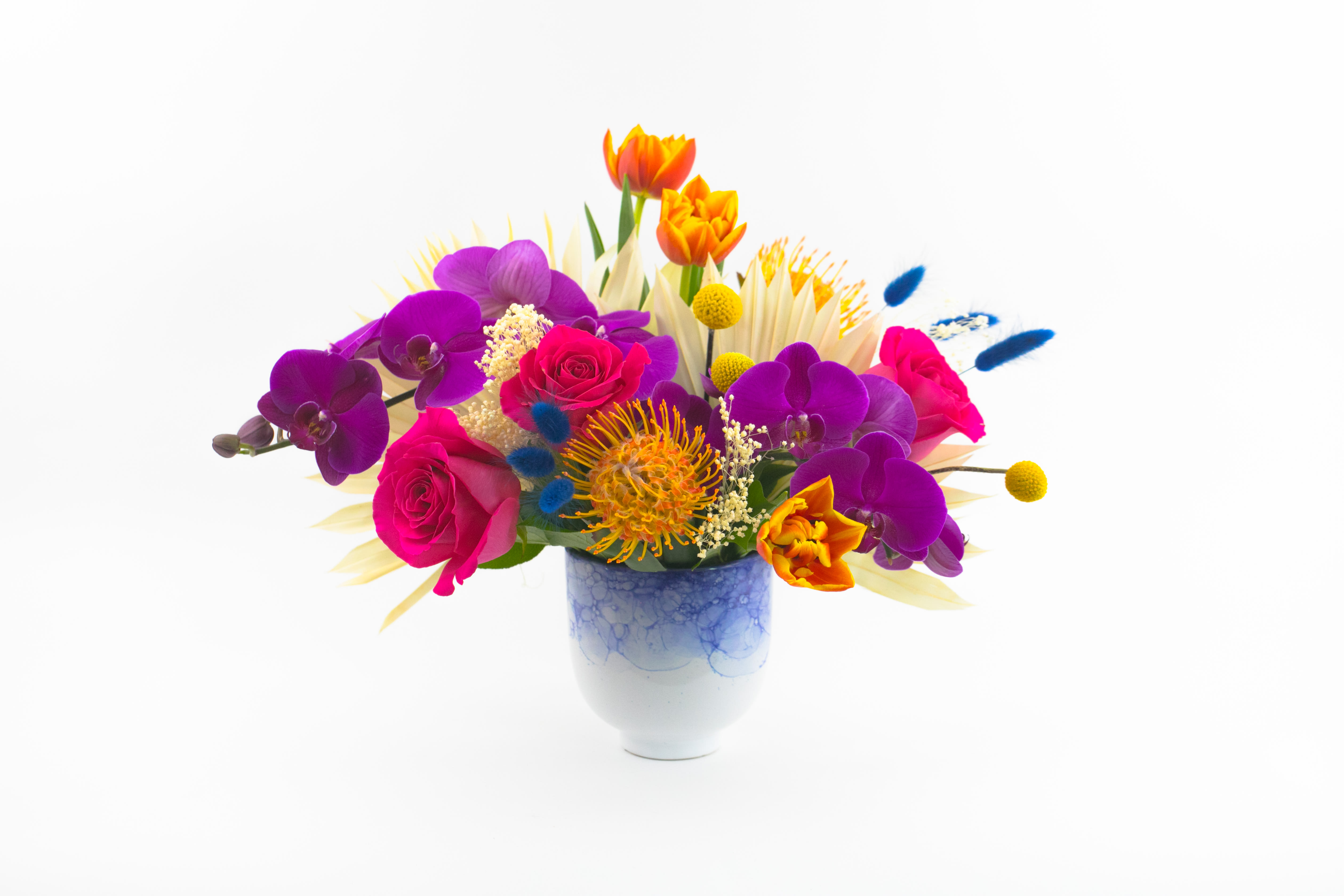 Exotic Attraction - QUIRKY FLOWER VASE ARRANGEMENT  Bright and contemporary. A beautifully designed vase of blooms featuring stunning fuchsia purple phalaenopsis orchid blooms, big hot pink ‘Pink Floyd’ roses, native yellow pincushion proteas and interesting grasses. Designed in a cute and quirky vase, this unique flower arrangement rocks! These flowers are perfect for arty and creative folk with a contemporary style.  Send this colorful vase of flowers to celebrate the birth of a new baby or wish someone a special happy birthday. Deliver to a client or colleague in recognition of an achievement or as a thank you.  Why not add a greeting card or quality handmade candle to match the occasion or personality?  Exotic Attraction features: ·        Bleached fan palm ·        Stunning purple phalaenopsis orchids ·        Yellow pincushion proteas ·        Hot pink Pink Floyd roses ·        Orange/yellow double tulips ·        Blue Bunny Tail Grass ·        Bleached grass sprigs ·        Craspedia (or ‘Billy Buttons’) ·        Cute white and blue ceramic vase   SIZE: The STANDARD flower arrangement is approximately 14” height and 14” width.   ALLERGY ADVICE: This design has low-moderate pollen and a low fragrance.   FAQ Q: What time will my flowers arrive and what if no-one is there to accept them? A: Excellent question! We’re unable to provide or guarantee specific delivery times. All our flowers are delivered anytime between 9-5pm, with exception to Sympathy designs for a funeral service. For full details around our delivery process and timings, please see our Terms and Conditions. Q: What days do you deliver flowers? A: All our flowers are available for San Francisco and Daly City delivery seven days a week, between 9am-5pm. Q: May I order for the same day delivery? A: Yes, same day deliveries are available if you place an order before 1pm. Q: May I request to change the design a bit? A: Yes, please make a note in the SPECIAL INSTRUCTION box when you place your online order. We will do our best to follow them. Q: Will my flowers look exactly as what is pictured? A: Depending on the season, colors or shades may vary. Sometimes we need to substitute flowers due to seasonal availability, but they’re always substituted for the same or greater value.   Ready to bring floral beauty into someone’s world with our stunning flower delivery? Order this gift online now or give us a call on (415) 752-2060 if you have a question or special request.   