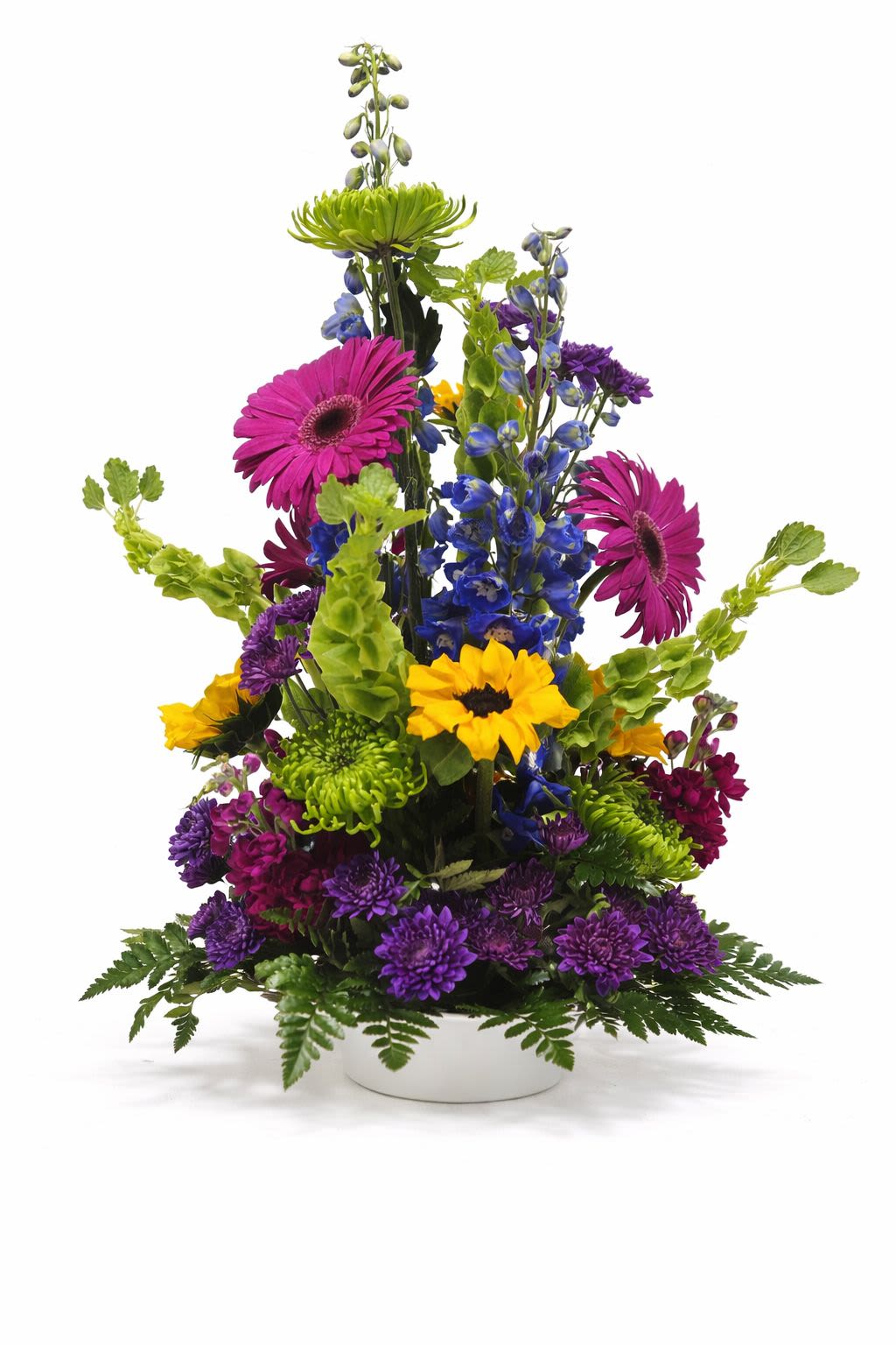 Fiesta Blooms - Bring bold color and joyful energy into any space with our vibrant Fiesta Bloom arrangement. Designed in a lush all-around (round) style, this piece looks beautiful from every angle—making it perfect for centerpieces, desks, or anywhere it can be fully appreciated.  This eye-catching design features a lively mix of bright sunflowers, hot pink gerbera daisies, rich purple mums, and striking blue delphinium, all accented with fresh seasonal greenery.  Artfully arranged in a low, modern container, its dynamic shape adds height and movement while maintaining a full, balanced look throughout. Whether you're sending a cheerful surprise or adding a pop of color to your home or office, Fiesta Bloom delivers a fun, fresh statement that feels like a celebration in full bloom.  SUBSTITUTION POLICY  Please note the bouquet pictured reflects our original design.  If the exact flowers or container in this arrangement are not available, our local florists will create a beautiful bouquet with the freshest available flowers.