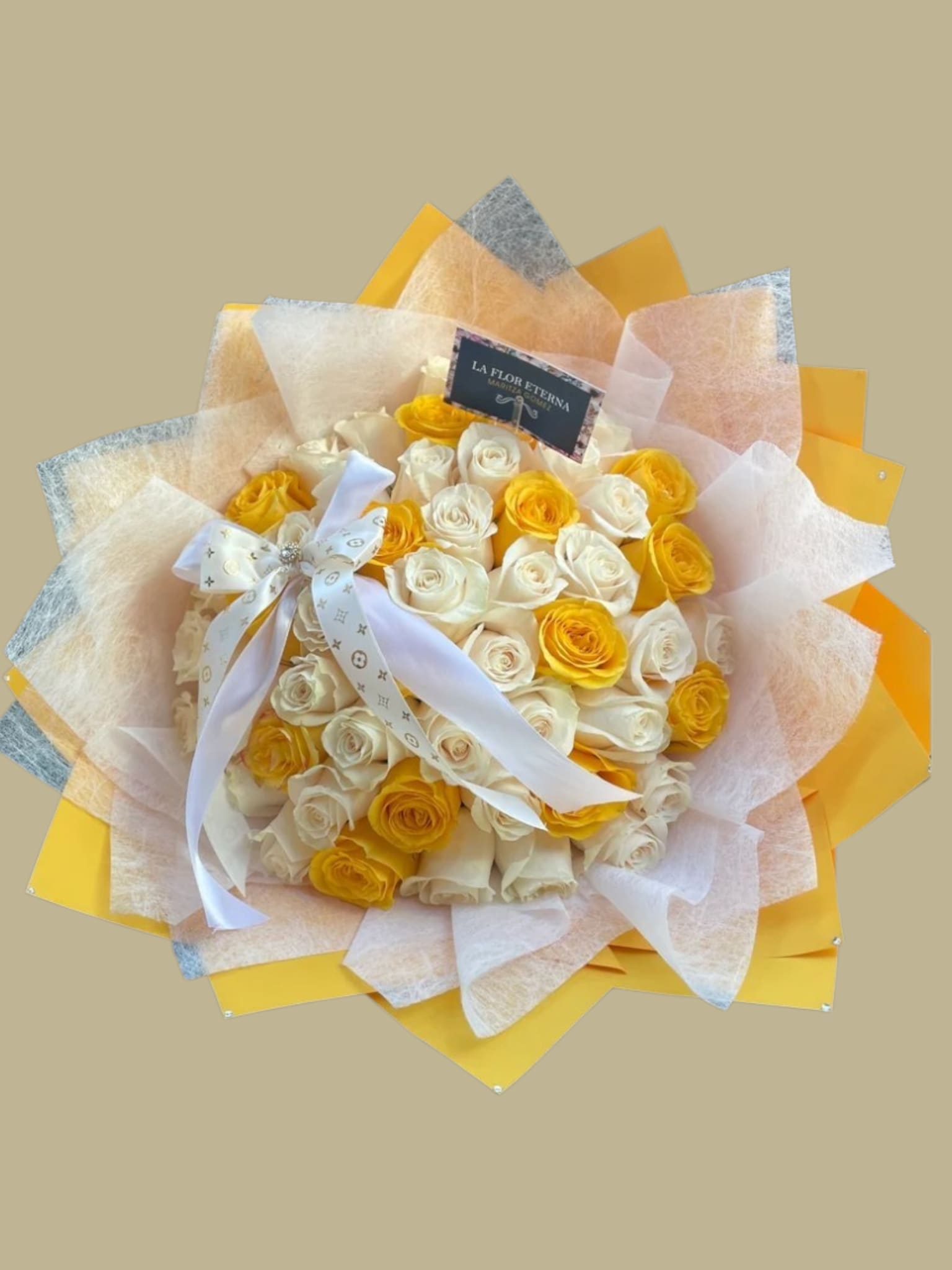 Golden Grace - A bright, domed bouquet of yellow and cream roses that reads like sunshine wrapped into a gift - perfect as a birthday bouquet, anniversary roses, or a romantic surprise that says "you brighten my days." The close, rounded cluster of blooms creates an immediate visual impact so the person you're thinking of will feel noticed and celebrated the moment they see it.  Carefully staged layers of yellow, white, and soft gray wrapping and tissue fan out around the roses for a starburst effect, finished with a wide white printed ribbon tied in a large bow and a brand card for a polished presentation. No-fuss care: keep in fresh water, trim stems every few days, and avoid direct heat to extend vase life. Sized to feel substantial without overwhelming - an elegant choice for celebrations, thank-yous, or simply letting someone know they matter.