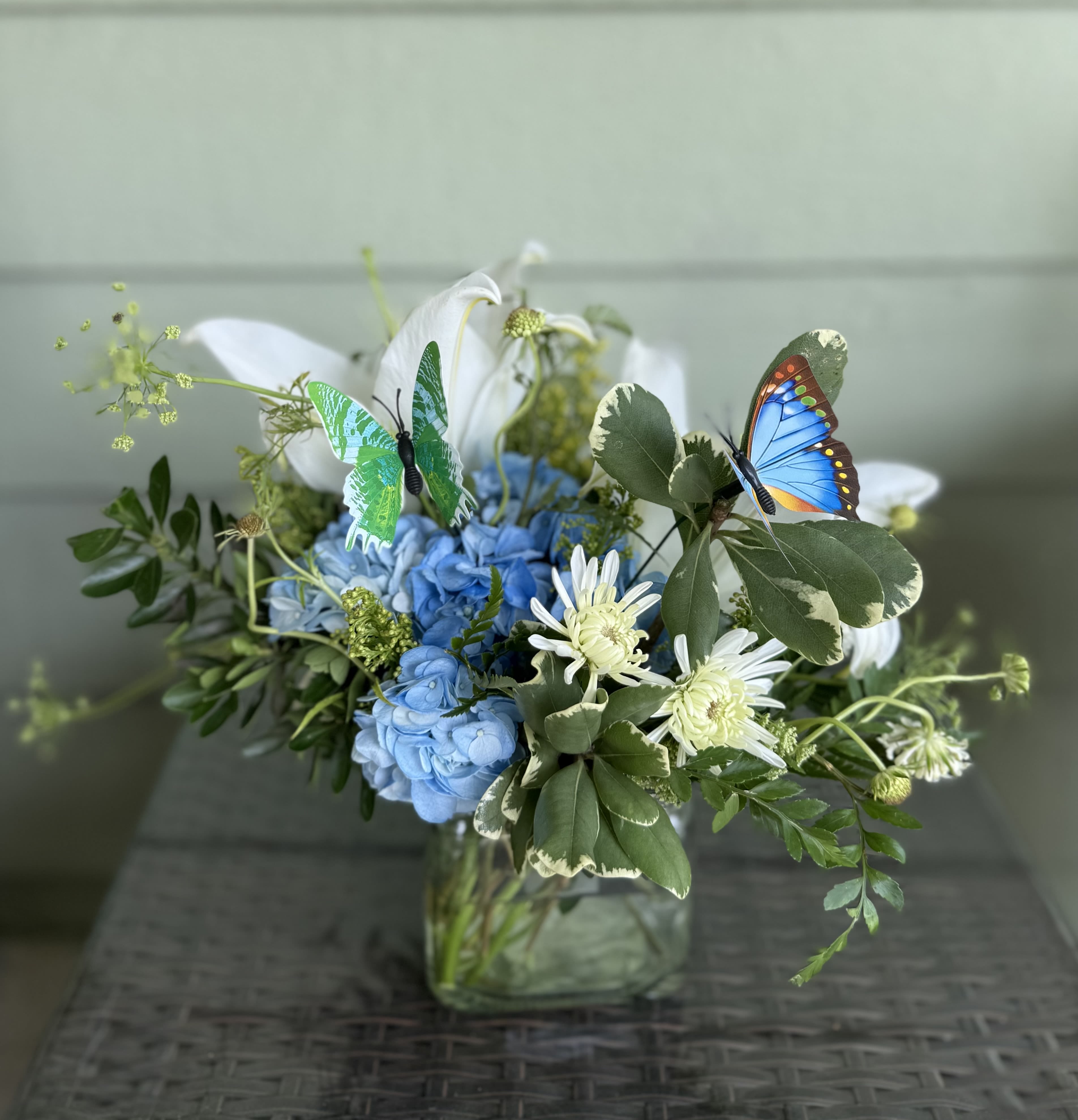 New Beginnings – Butterfly Floral Arrangement - Celebrate fresh starts and hopeful horizons with our New Beginnings arrangement — a serene blend of blue hydrangeas and elegant white lilies, thoughtfully arranged in a modern square vase. Delicate butterfly accents flutter among the blooms, symbolizing transformation, peace, and the beauty of change. Perfect for welcoming a new baby, sending encouragement, or marking any meaningful milestone, this arrangement brings calm, grace, and inspiration to any space.