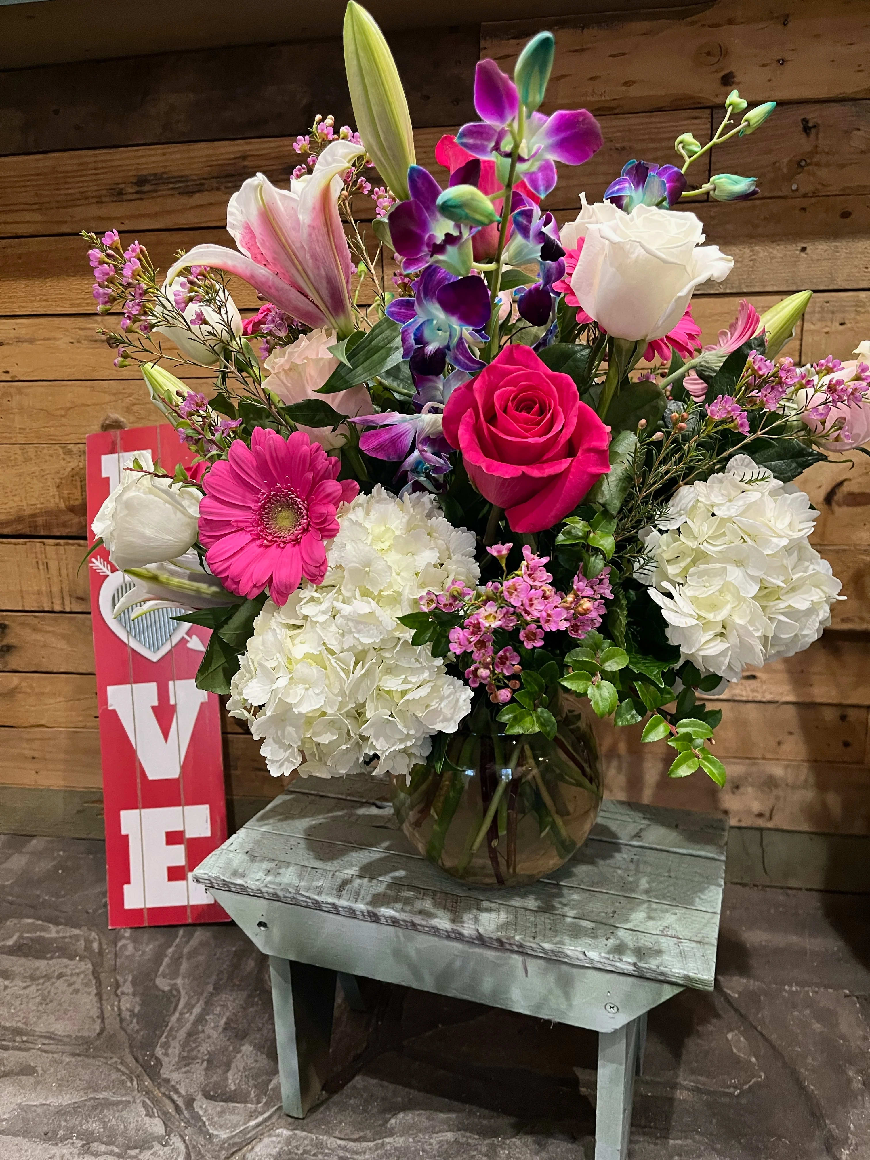 Mixed arrangement of pink, white, and purple flowers in a glass bowl vase on a rustic wooden bench