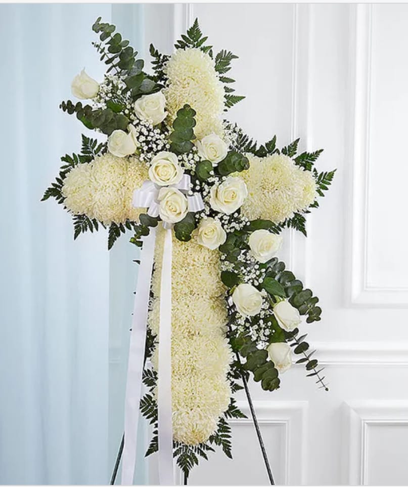Peace &amp; Prayers™ White Standing Cross - A large standing cross crafted from densely packed white chrysanthemums and other white blooms creates a soft, cushiony surface that reads as both reverent and serene-perfect for sympathy, memorial, or funeral services. A diagonal spray of white roses and delicate filler flowers adds a graceful focal point while darker foliage and rounded greenery trace the cross's edges, giving the arrangement clear structure and a natural, garden-like silhouette.  Mounted on a sturdy metal easel stand and finished with a white ribbon bow and long ribbon tails, this piece conveys quiet respect and heartfelt remembrance for the person you're honoring. The generous scale and plush texture make it meaningful at a funeral chapel or graveside; no special care is required beyond standard florist delivery and placement on the included easel.