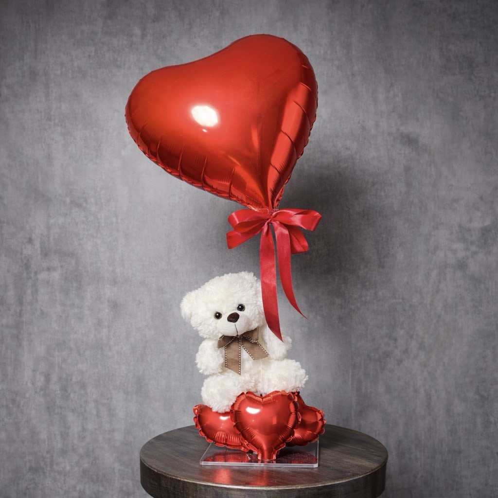 White teddy bear with bow sitting among red heart balloons and a large red heart balloon above.